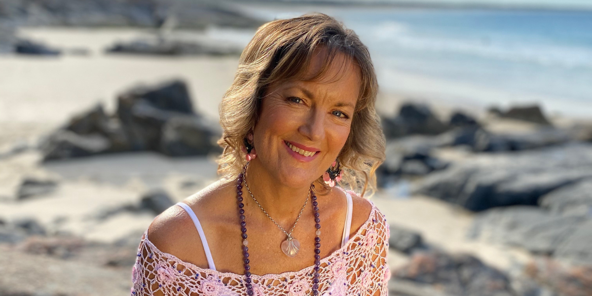 Smiling woman with curly hair sitting on rocky beach, wearing a pink top and jewelry