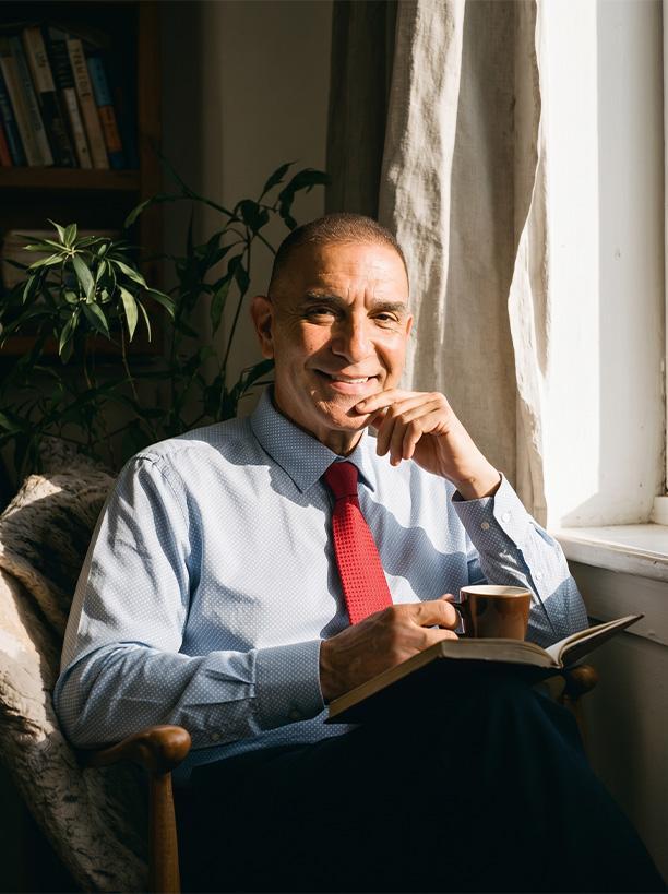 Man in a suit sitting by a window, holding a book and a coffee cup
