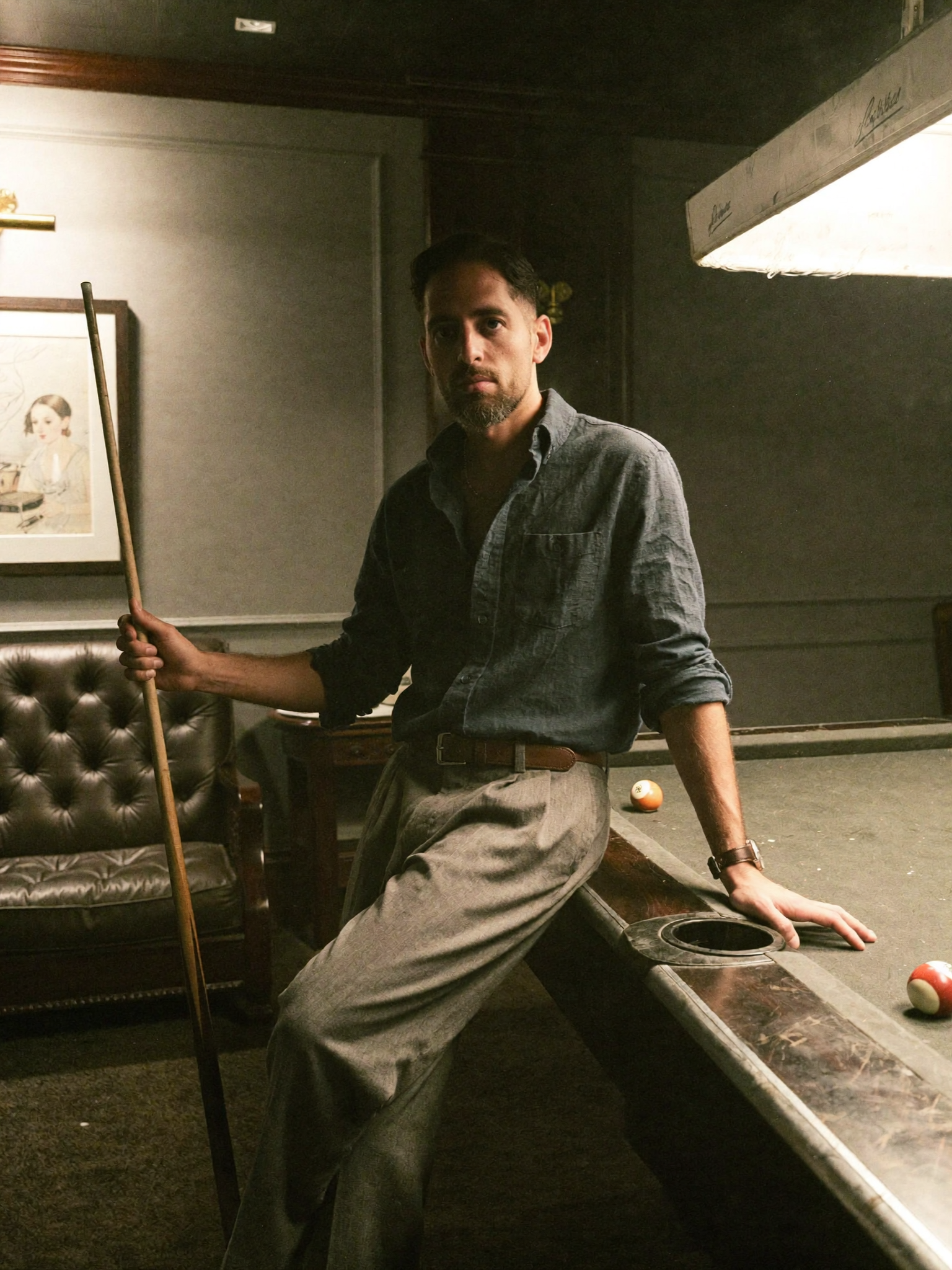 A man in a gray shirt leans on a pool table in a dimly lit room