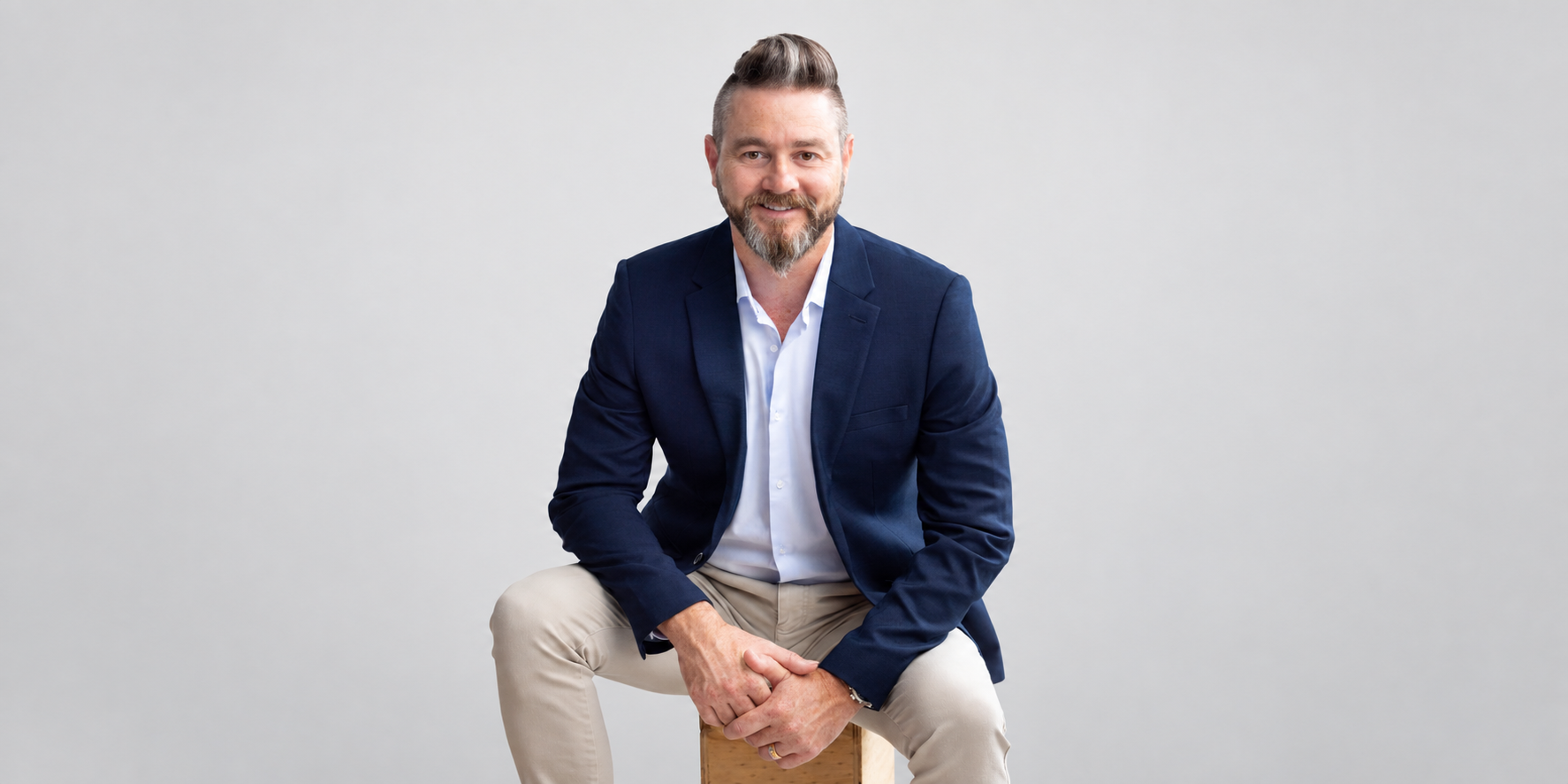 Man in a blazer and beige pants sitting on a stool against a gray background