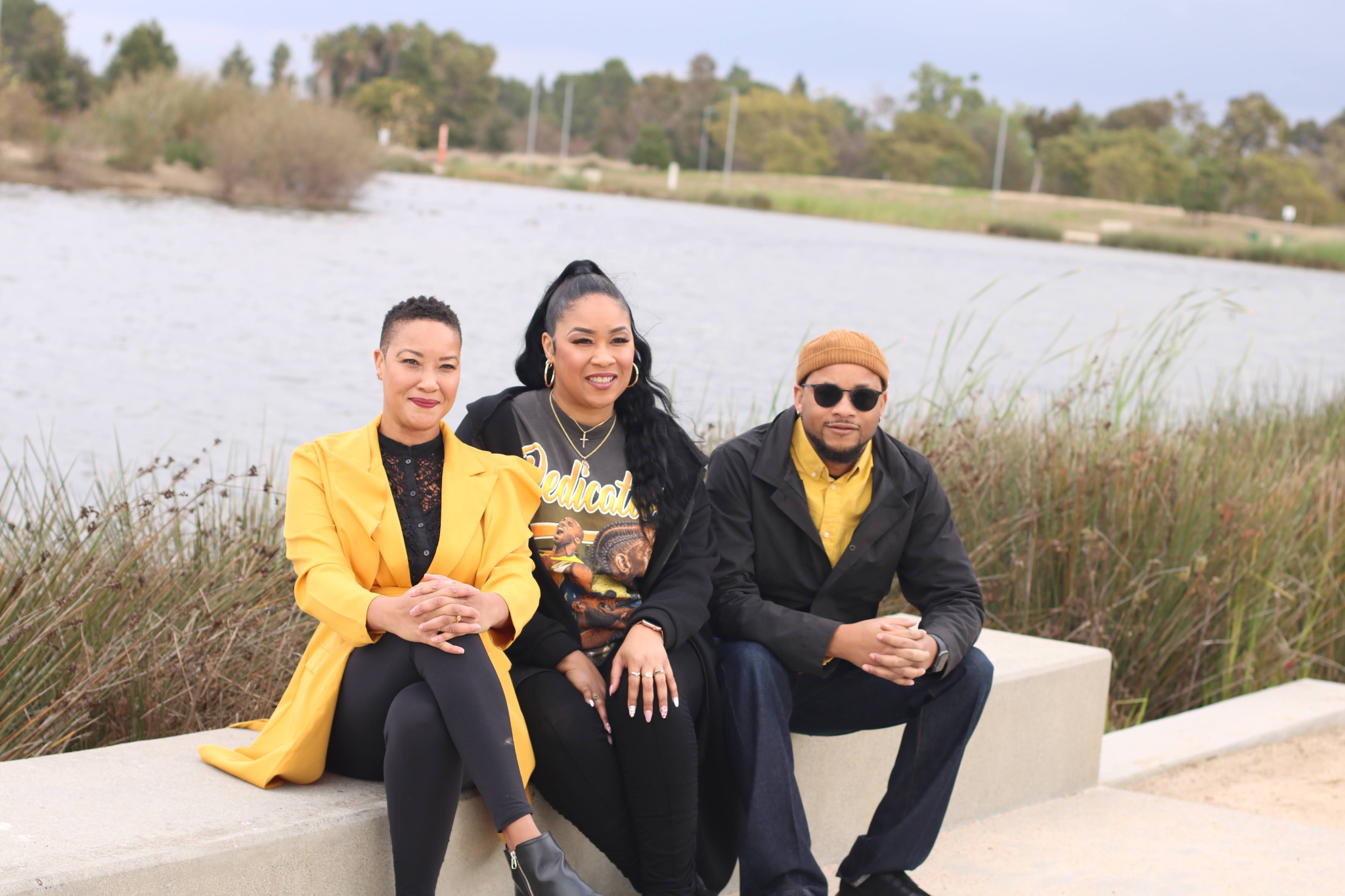 Three people sitting by a lake, smiling, dressed in casual and stylish clothing