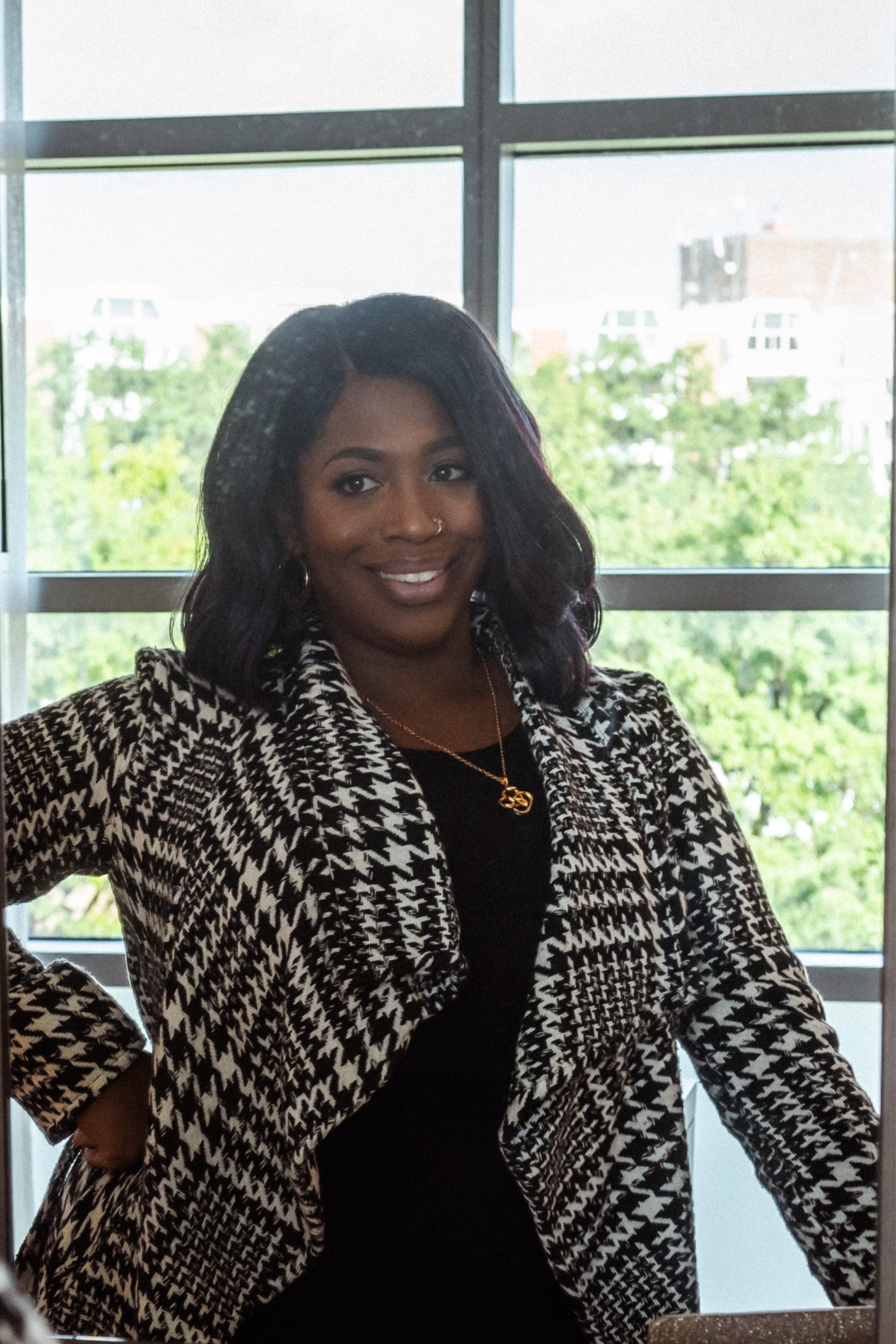 Smiling woman in a stylish houndstooth coat poses near a window with greenery outside