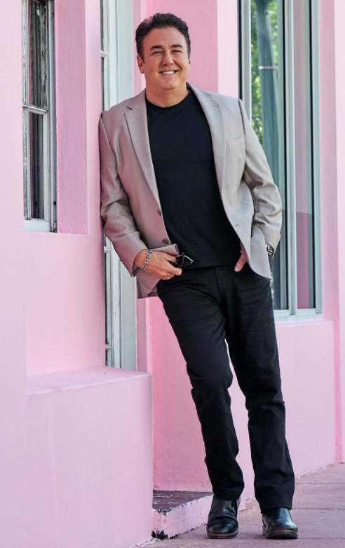 Man in a gray blazer and black shirt poses against a pink wall
