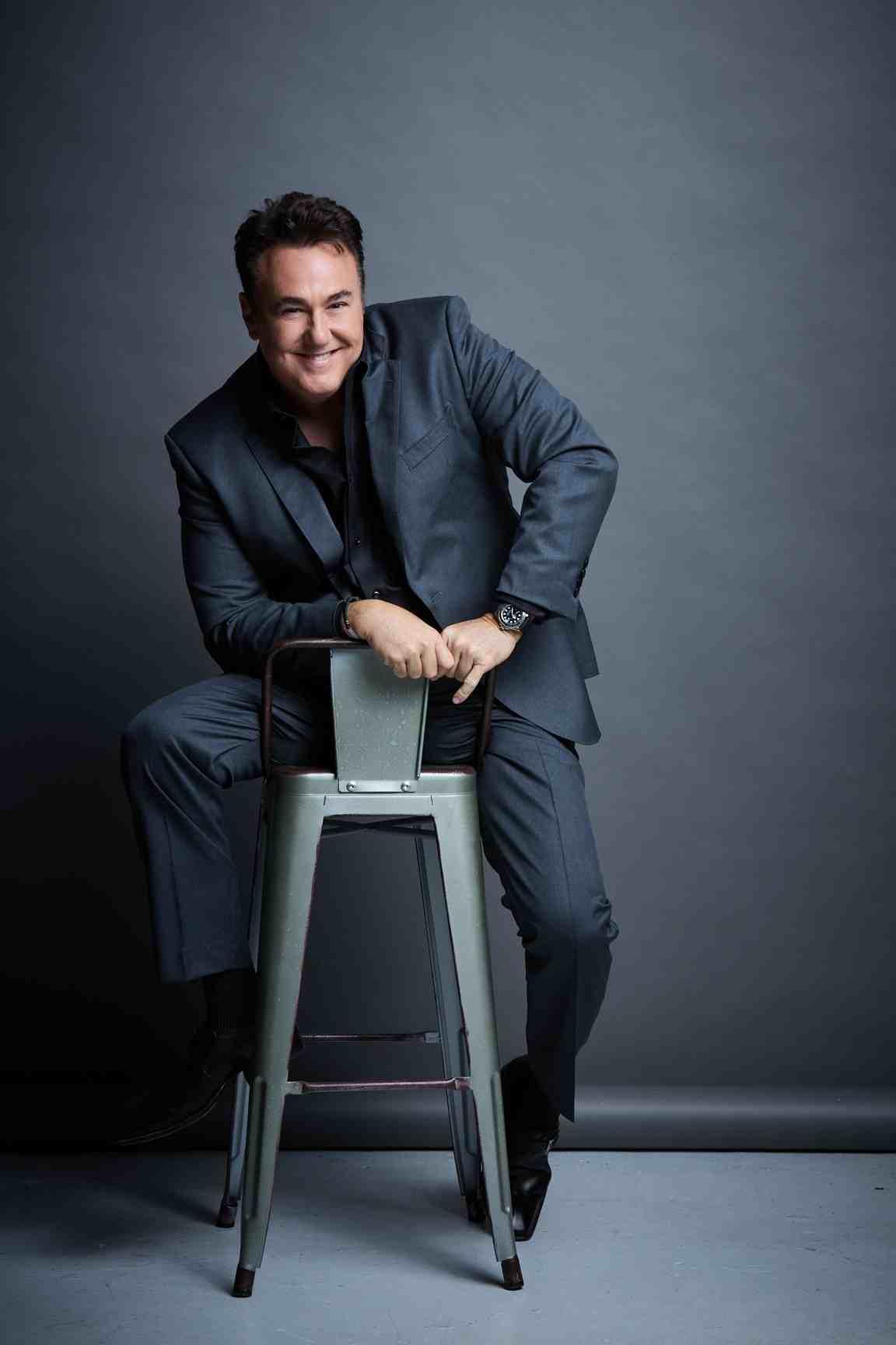 Man in a suit seated on a metal stool, smiling against a gray backdrop