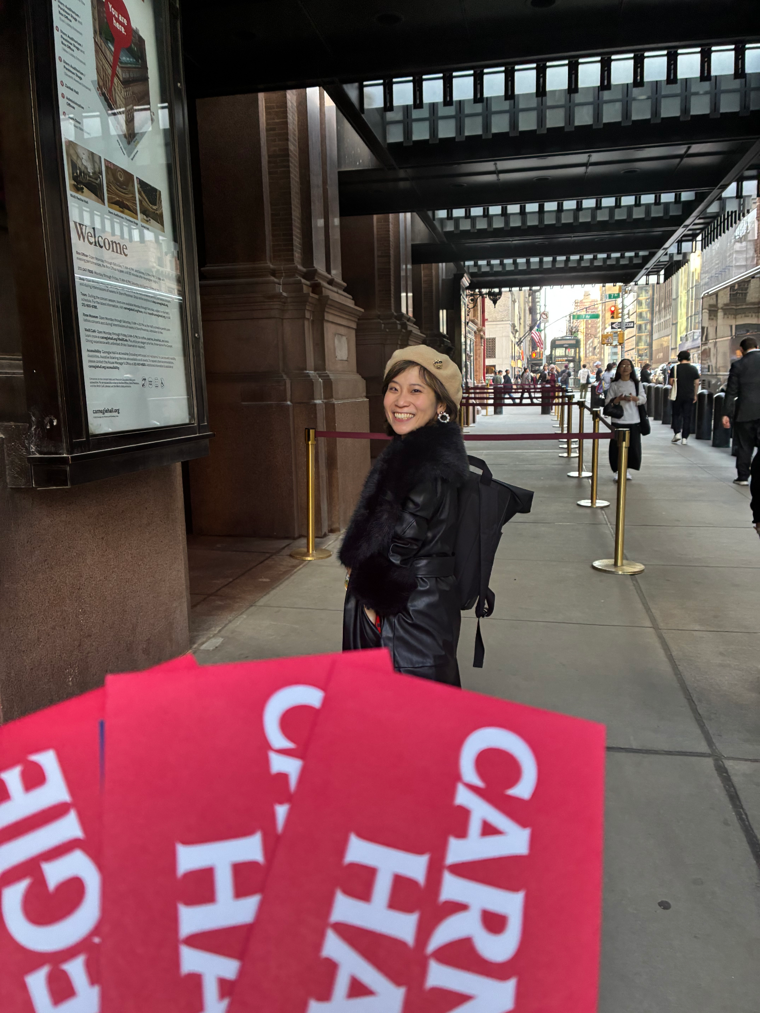 A woman smiling outside Carnegie Hall, holding tickets, with a bustling crowd.