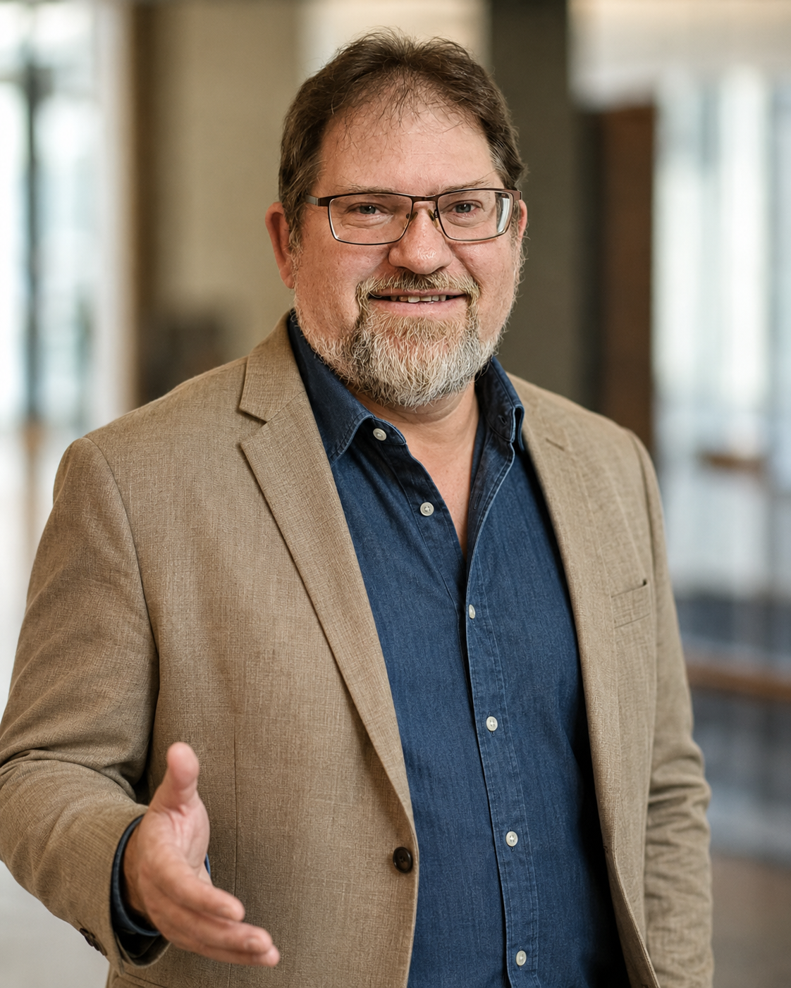 A smiling middle-aged man in a beige blazer and blue shirt gesturing with his hand