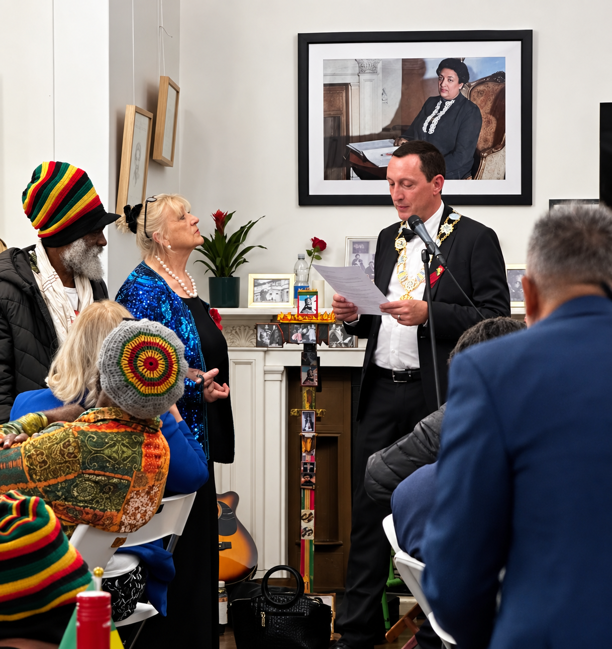 A man in formal attire reads while two people listen in a decorated room