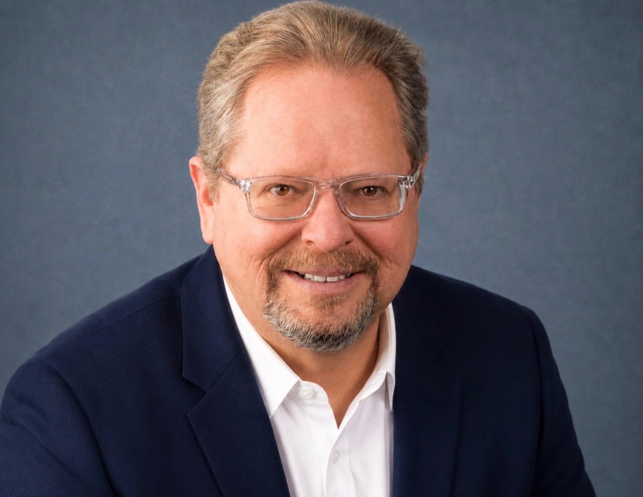 Smiling man with glasses wearing a blue blazer and white shirt against a blue background
