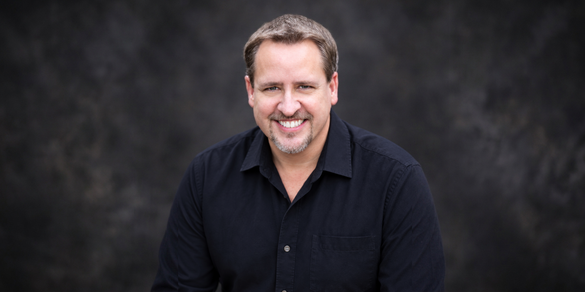 Smiling man in a black shirt against a dark textured background