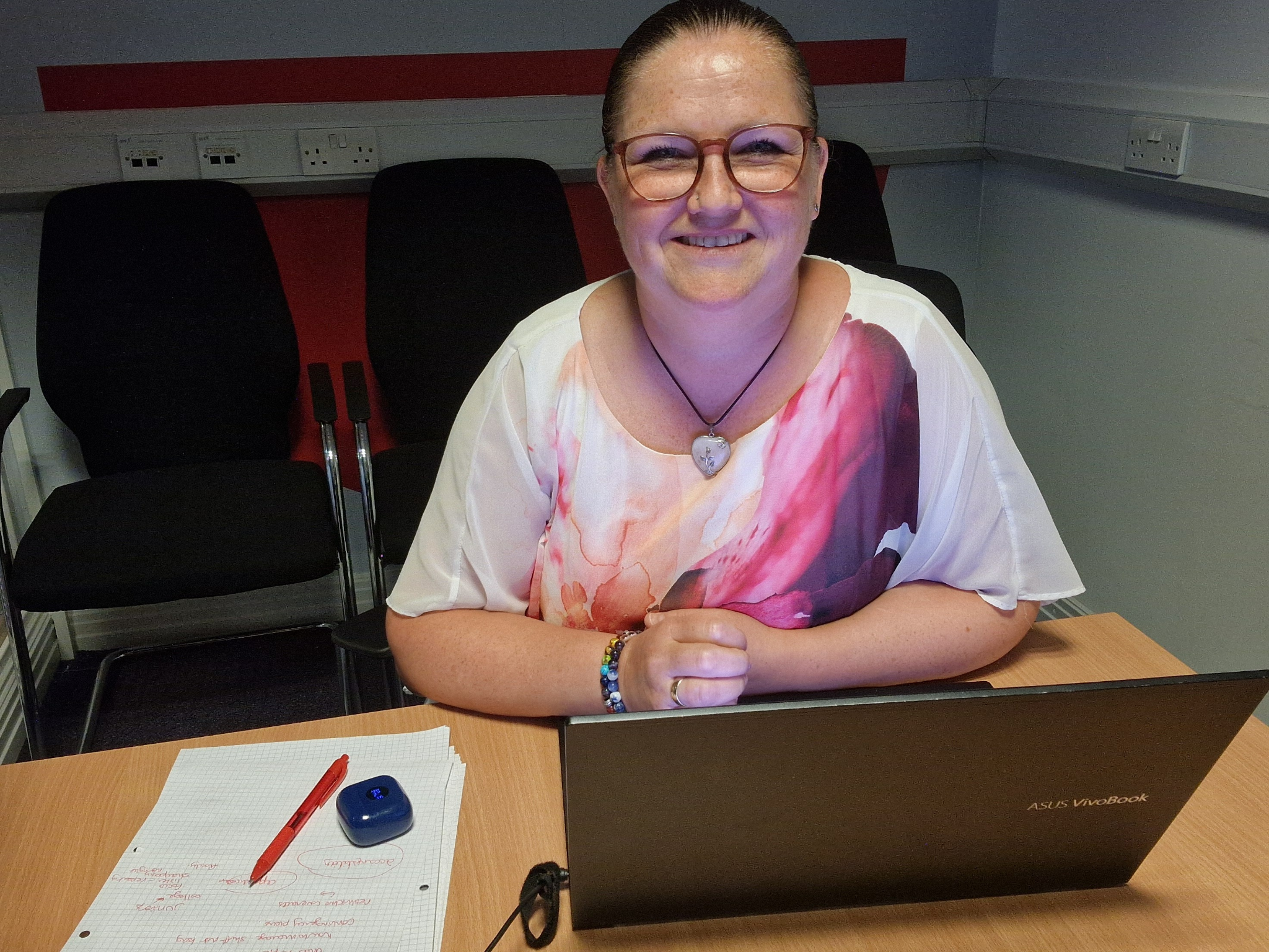 A woman with glasses smiles at a laptop in a meeting room.