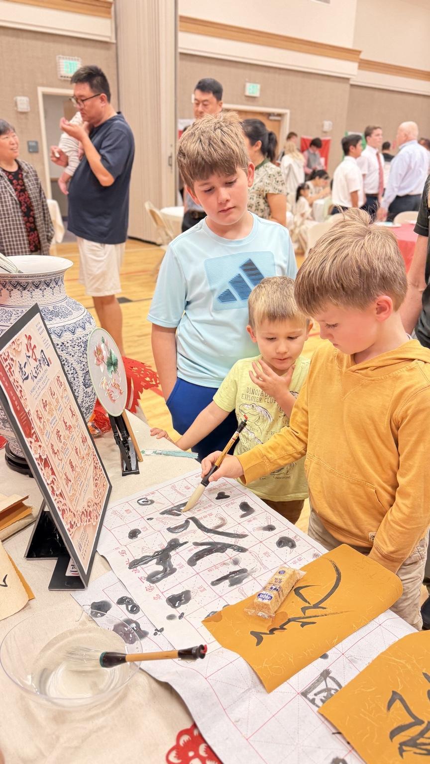 Children practicing Chinese calligraphy at a cultural event with adults in the background