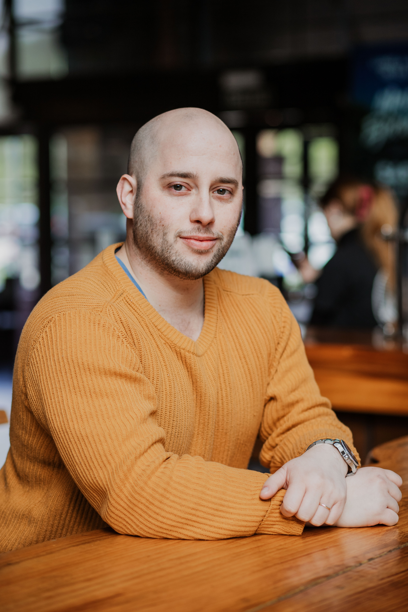 Man with a bald head wearing a mustard sweater seated at a wooden table