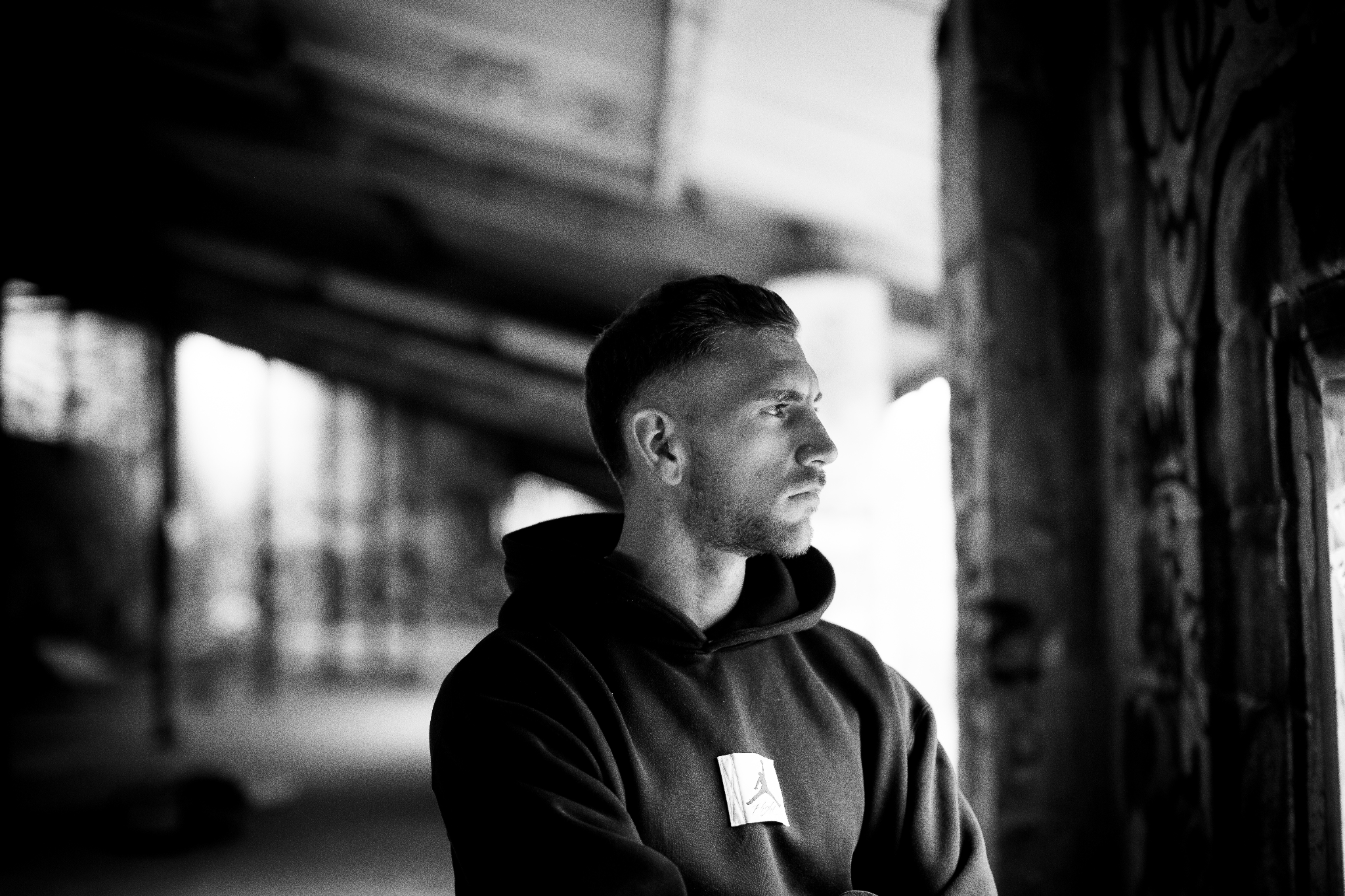 A man in a hoodie gazes thoughtfully out a window in a graffiti-covered space