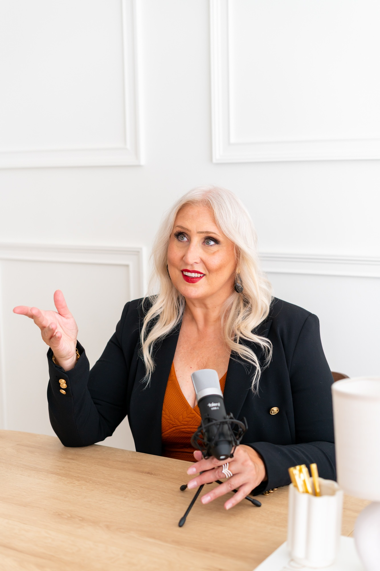 Woman with long blonde hair sits at a table, gesturing during a podcast recording