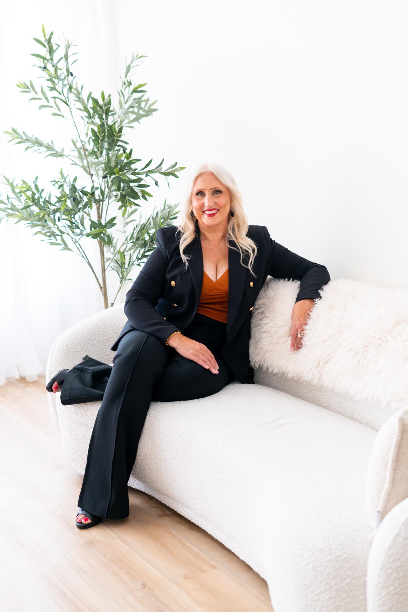 Woman in a stylish black suit sitting on a white sofa with a plant.