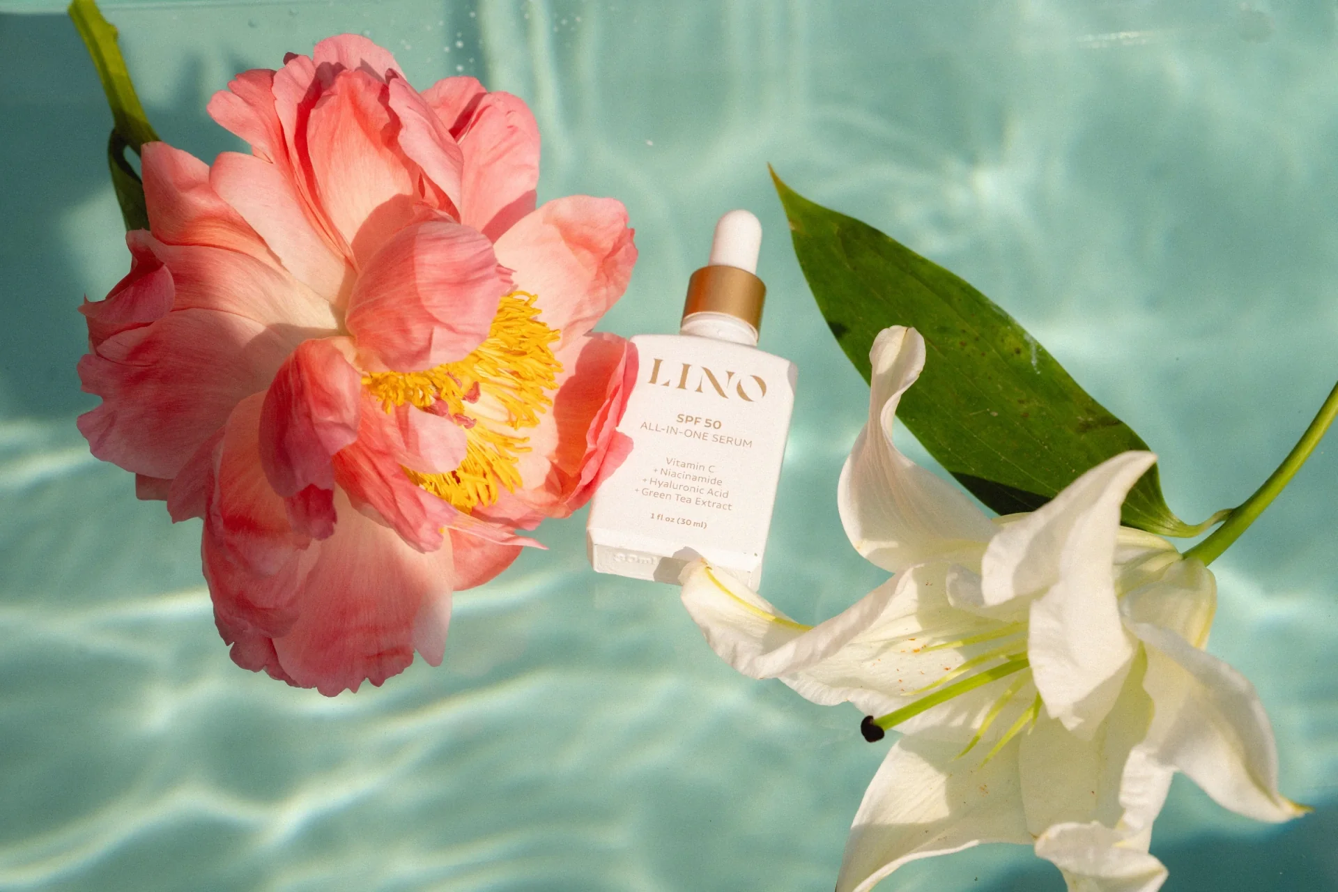 A pink flower, white lily, and a skincare serum float in clear water