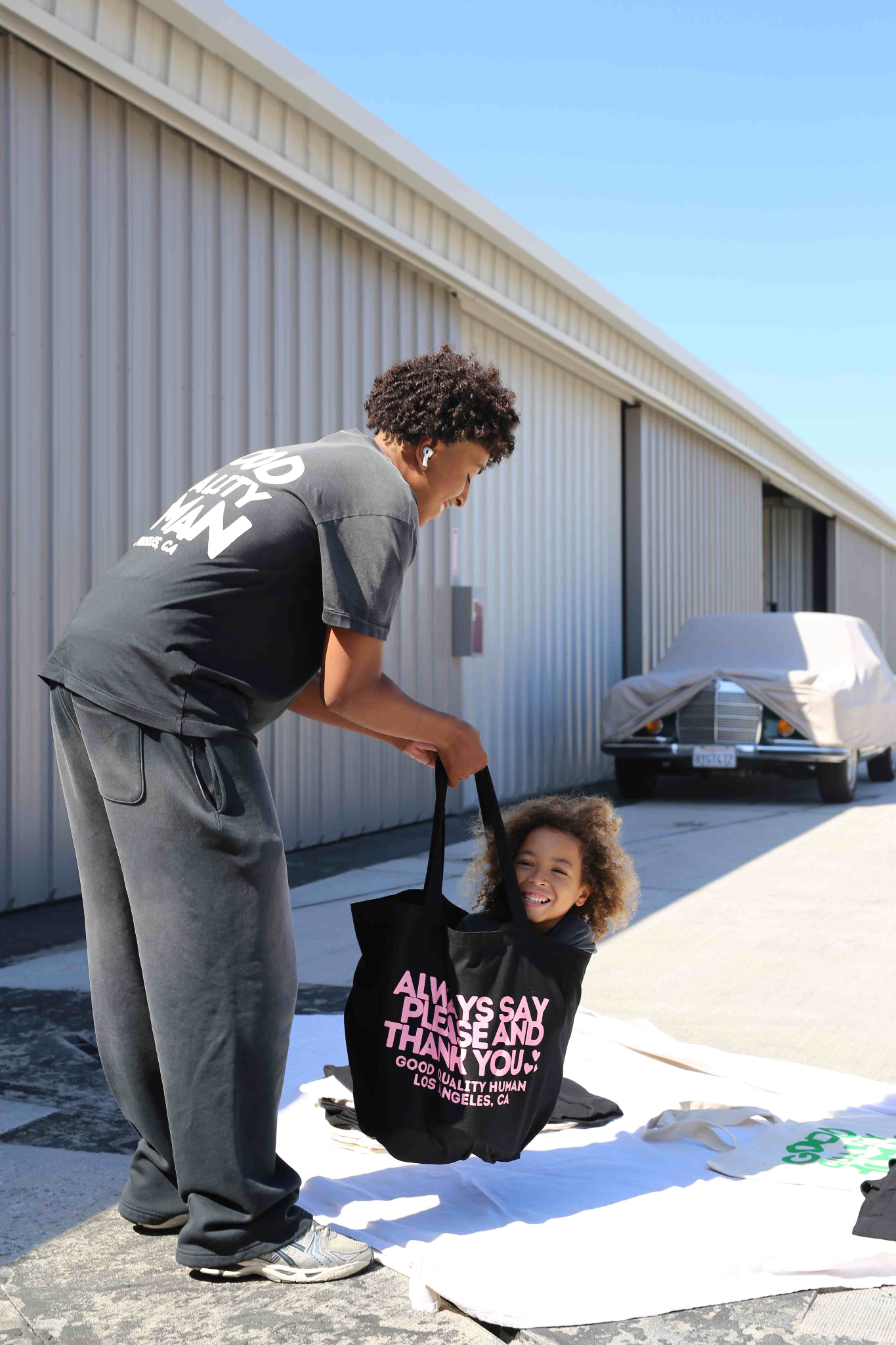 A child sits in a tote bag while an adult holds it outdoors.