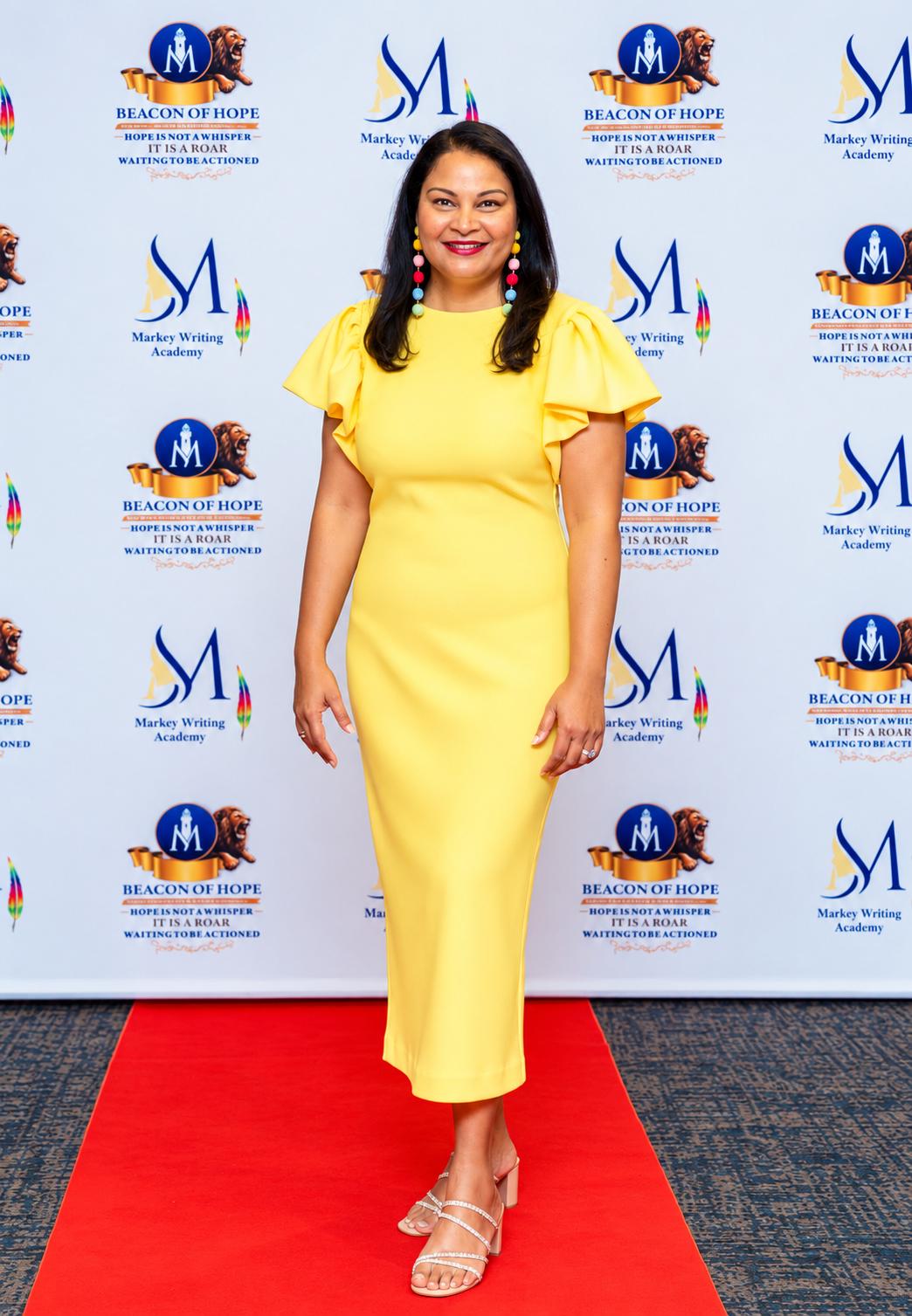 Woman in a yellow dress poses on a red carpet backdrop with logos