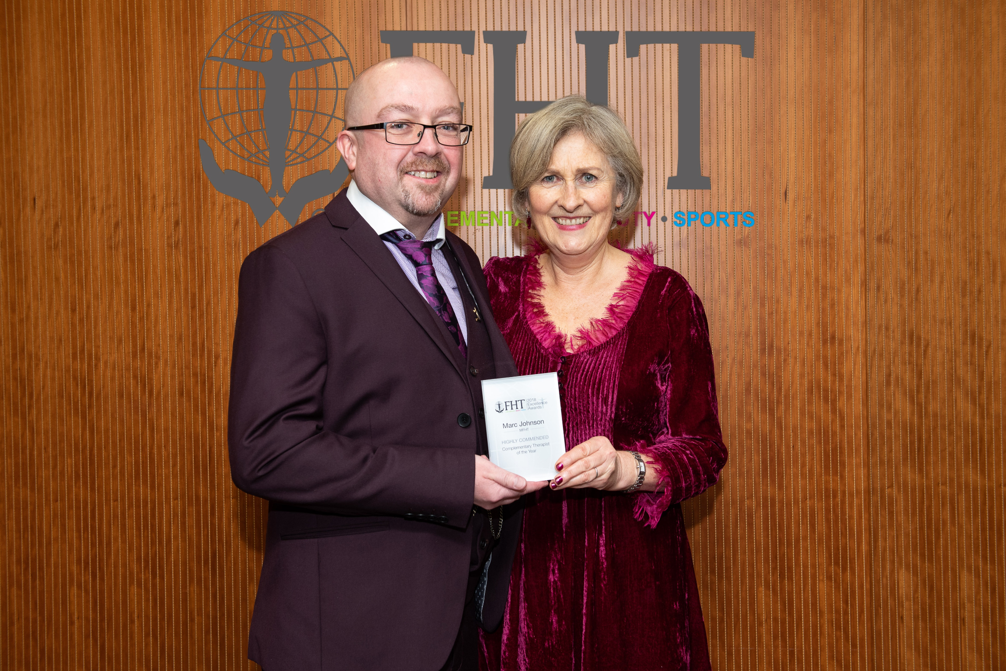 Two individuals pose together holding an award in front of a branded backdrop