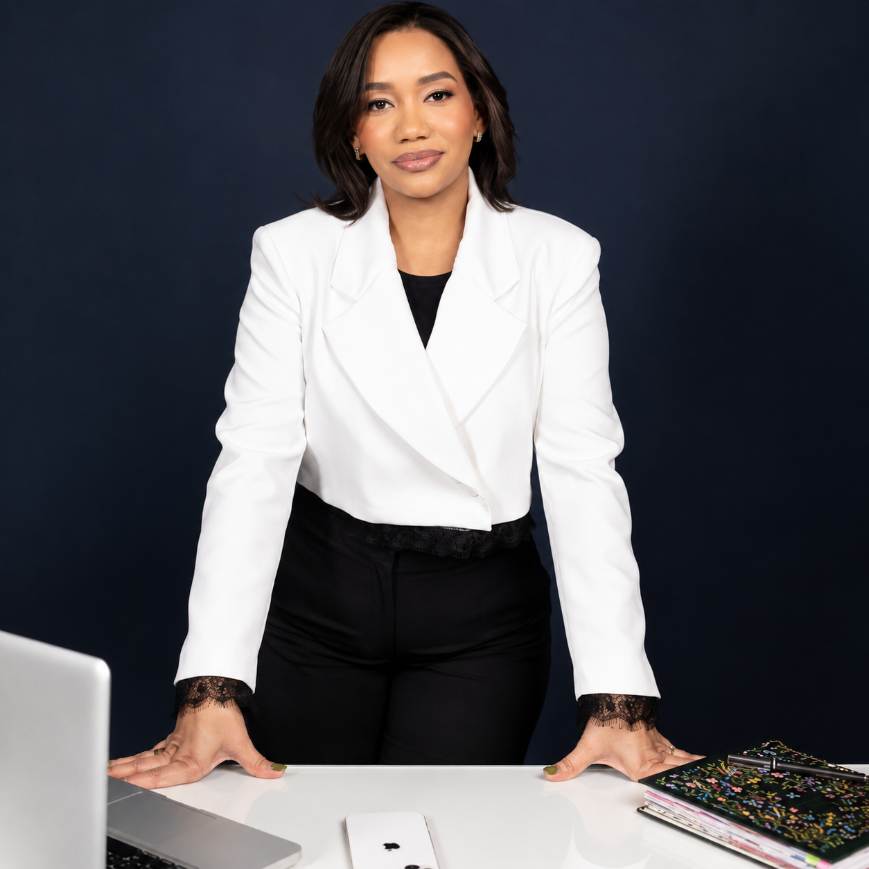 Professional woman in a white blazer stands at a desk with a laptop and notebooks