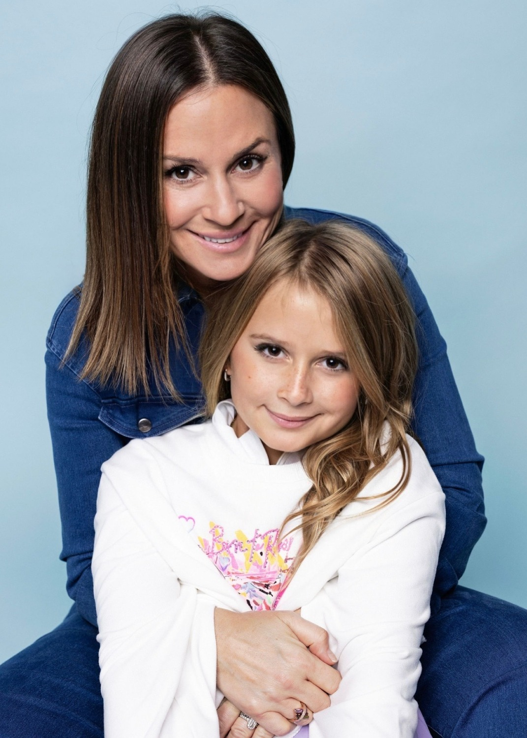 A woman and a girl pose together, both smiling in a light blue setting