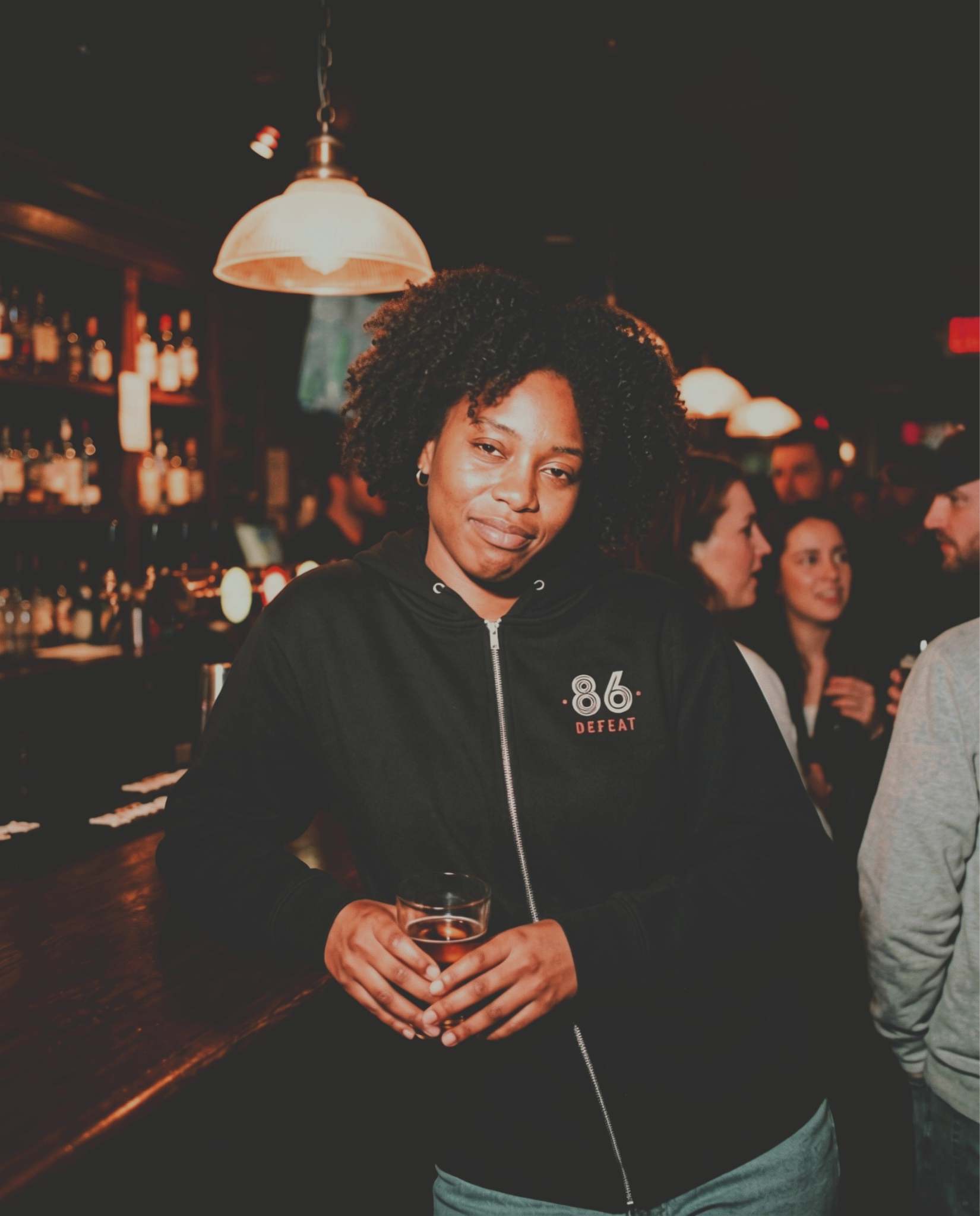 A woman with curly hair leans on a bar holding a drink in a dimly lit setting