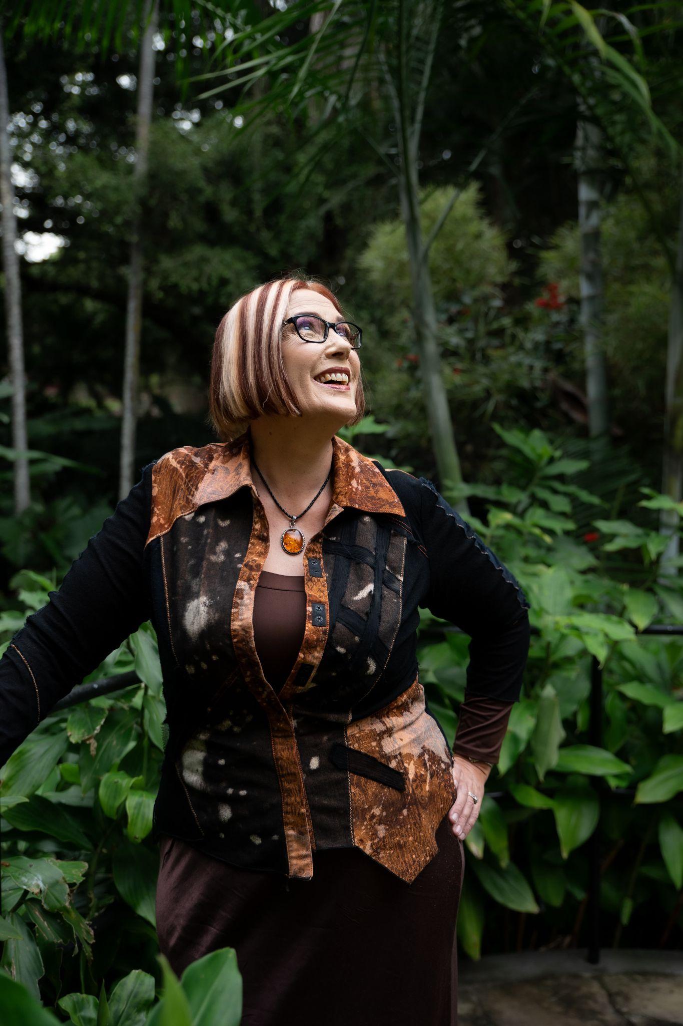 Woman with glasses and stylish hair poses in a lush, green garden setting