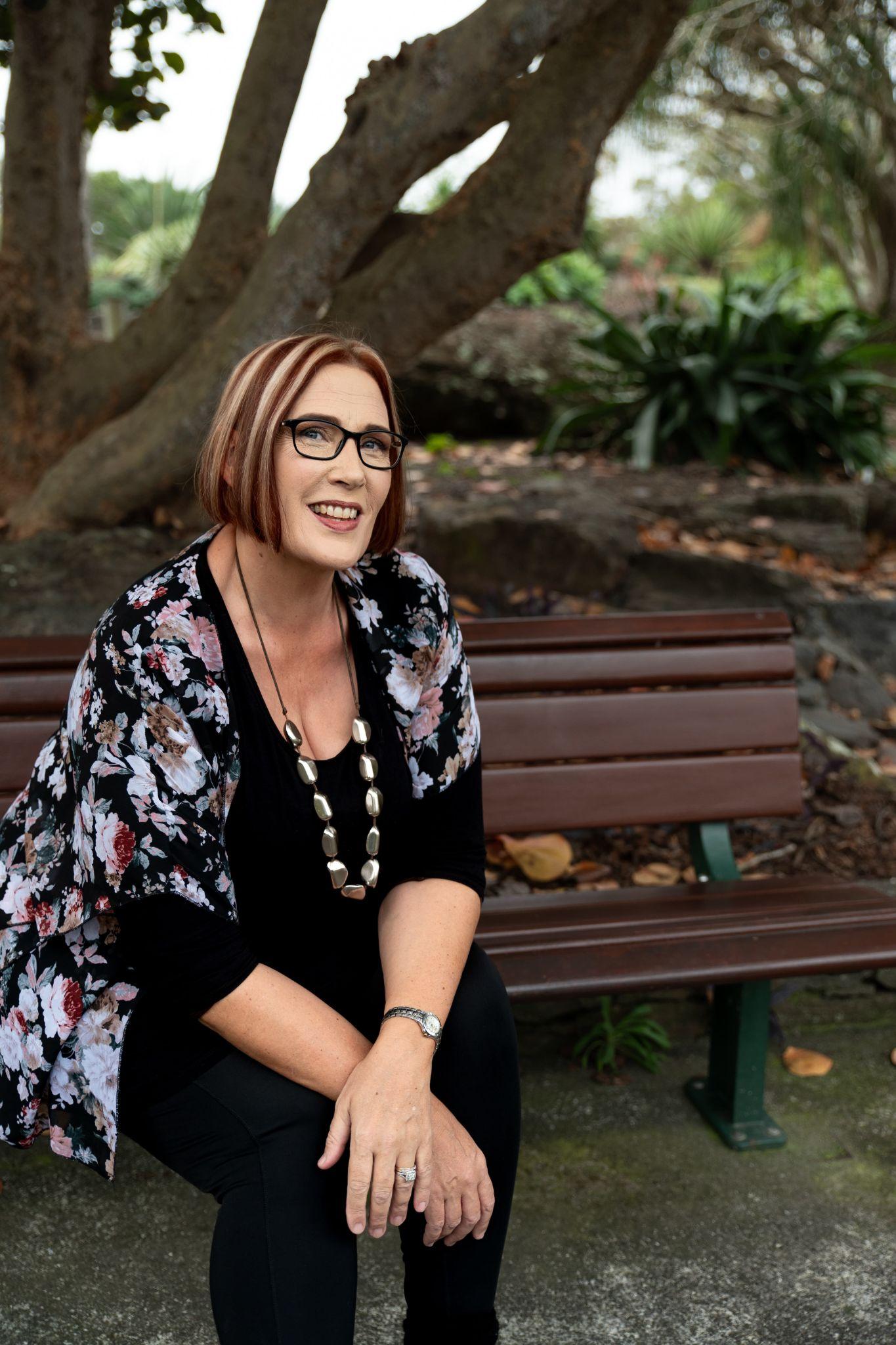 Woman with short hair and glasses sits on a park bench, smiling warmly.