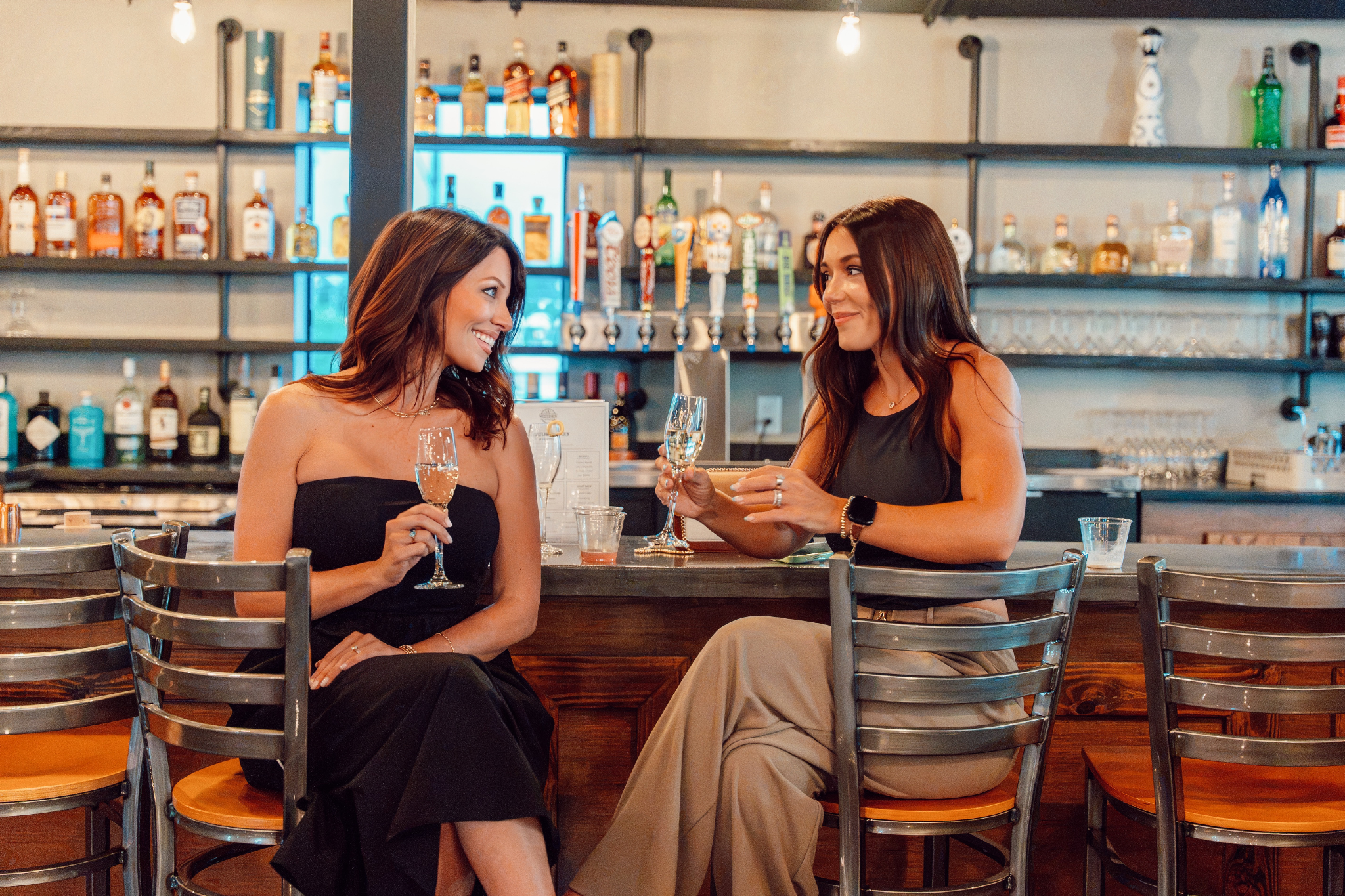 Two women enjoying drinks at a bar, smiling and chatting with a variety of bottles in the background