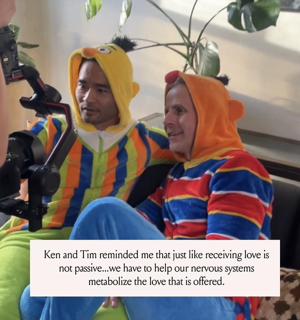 Two men in colorful onesies sitting together on a couch, smiling at the camera