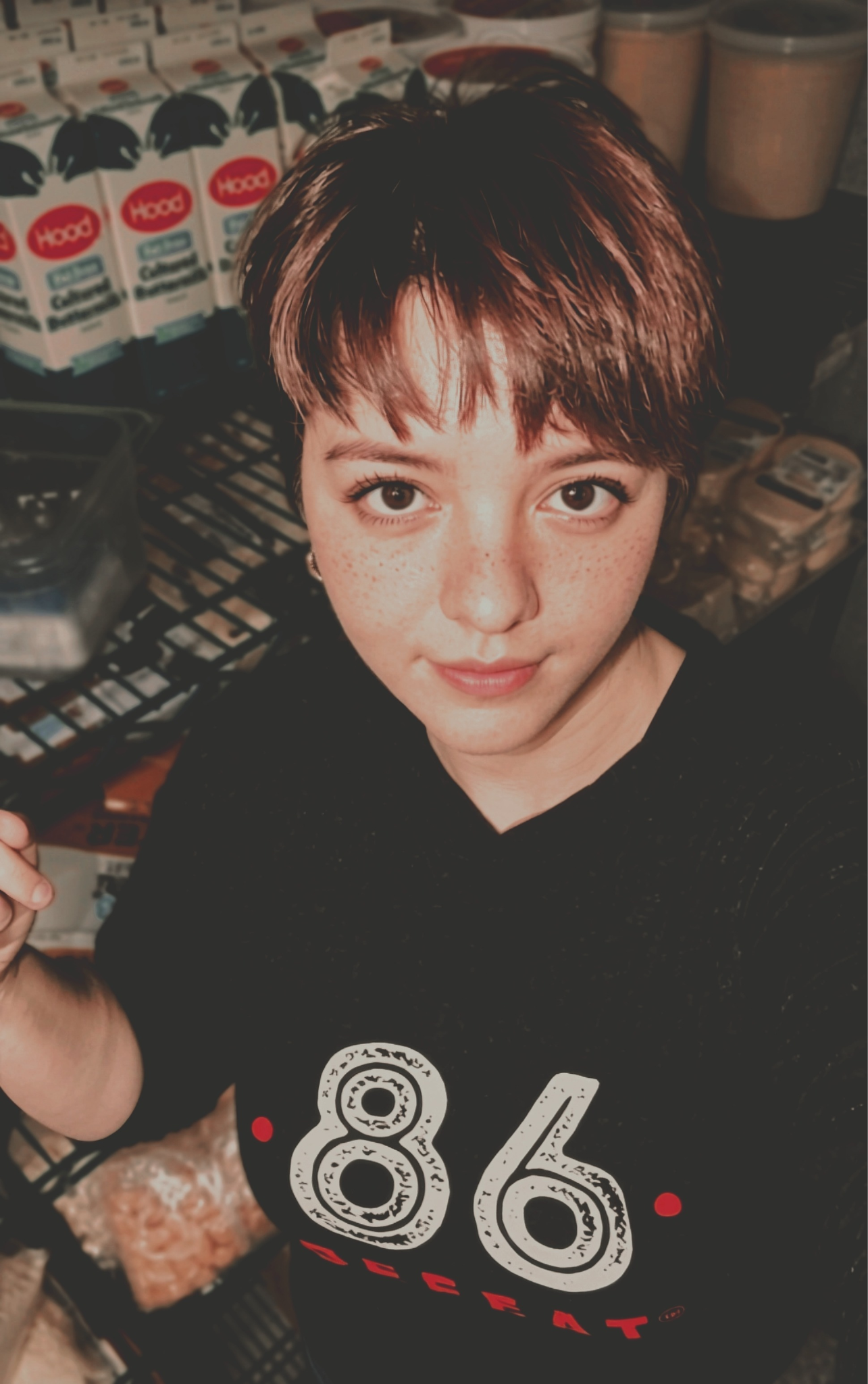 Person with short hair and freckles smiling in a storage area filled with food items