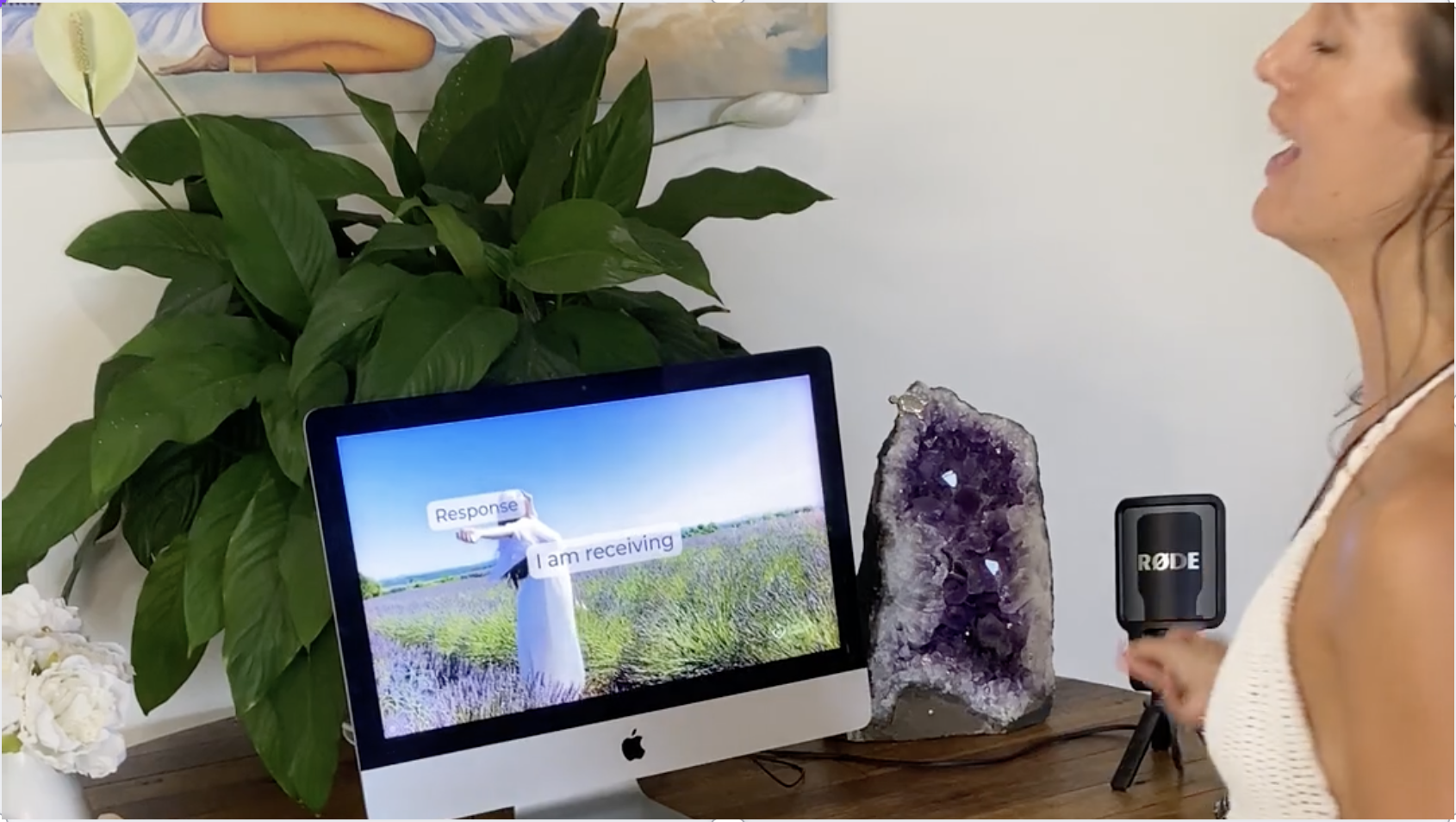 A woman speaks beside a computer displaying a serene nature image and a microphone