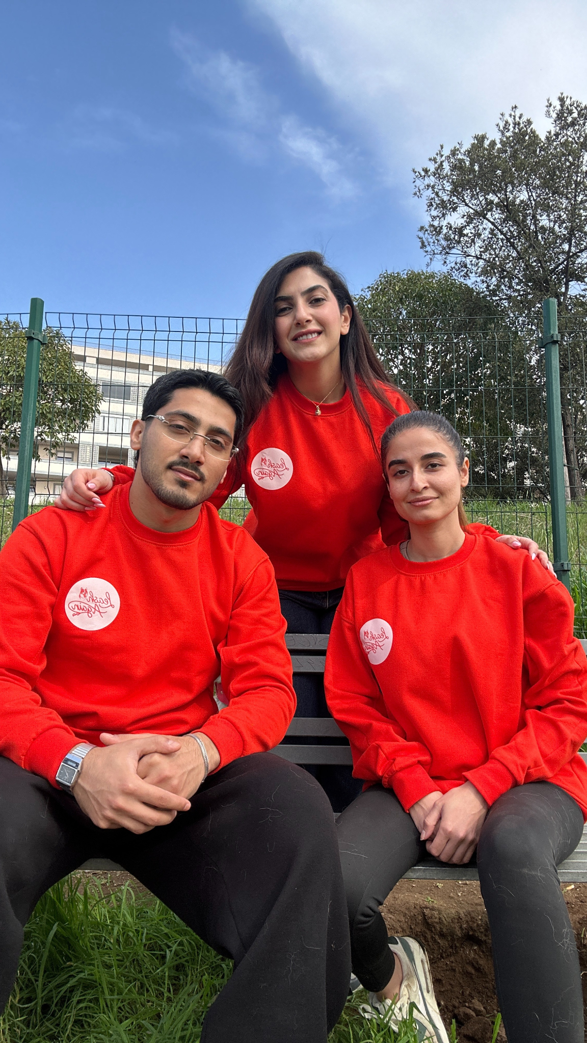 Three young adults in matching red sweatshirts pose together outdoors against a blue sky