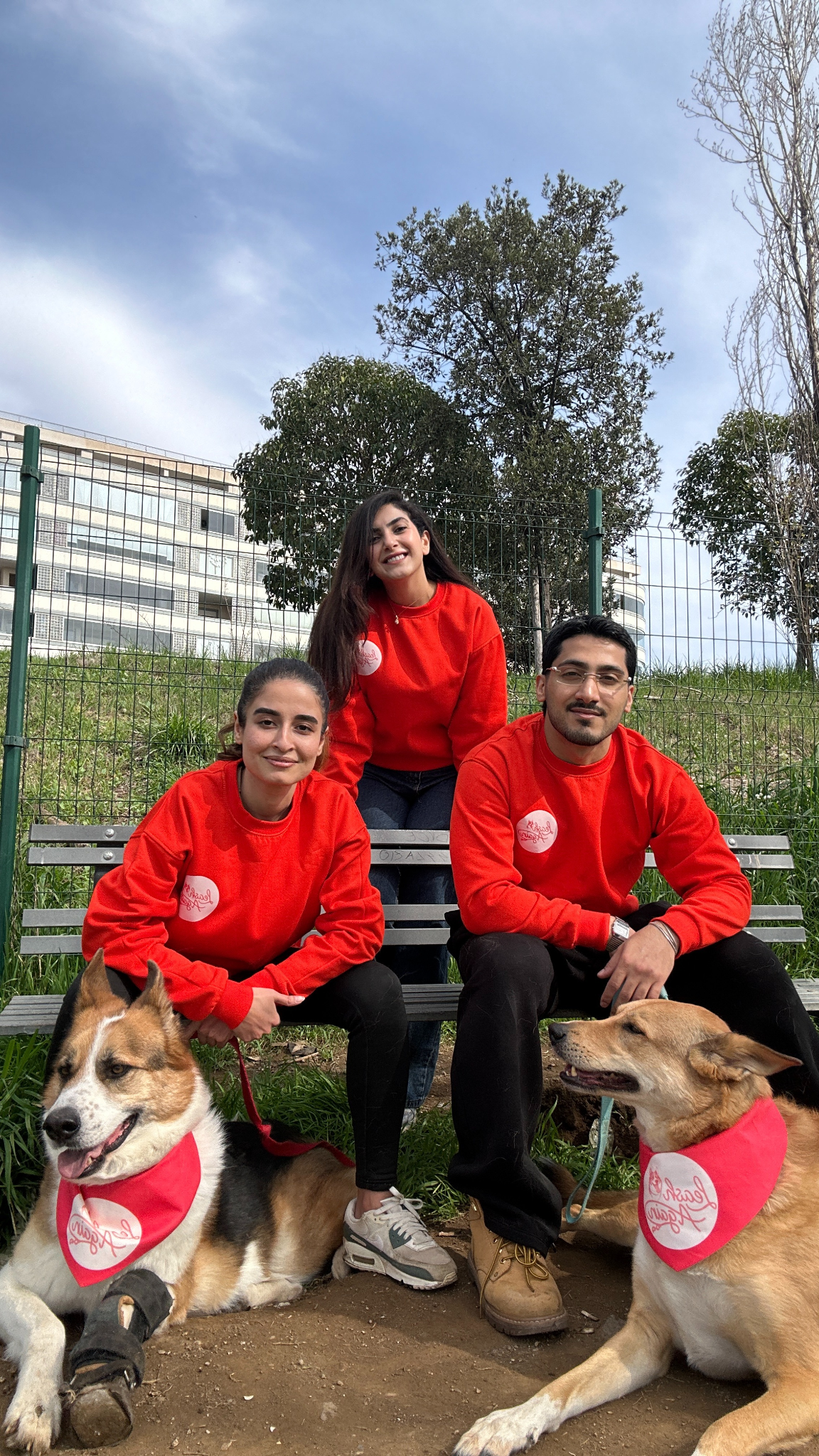 Three people in red sweatshirts sit on a bench with two dogs in a park