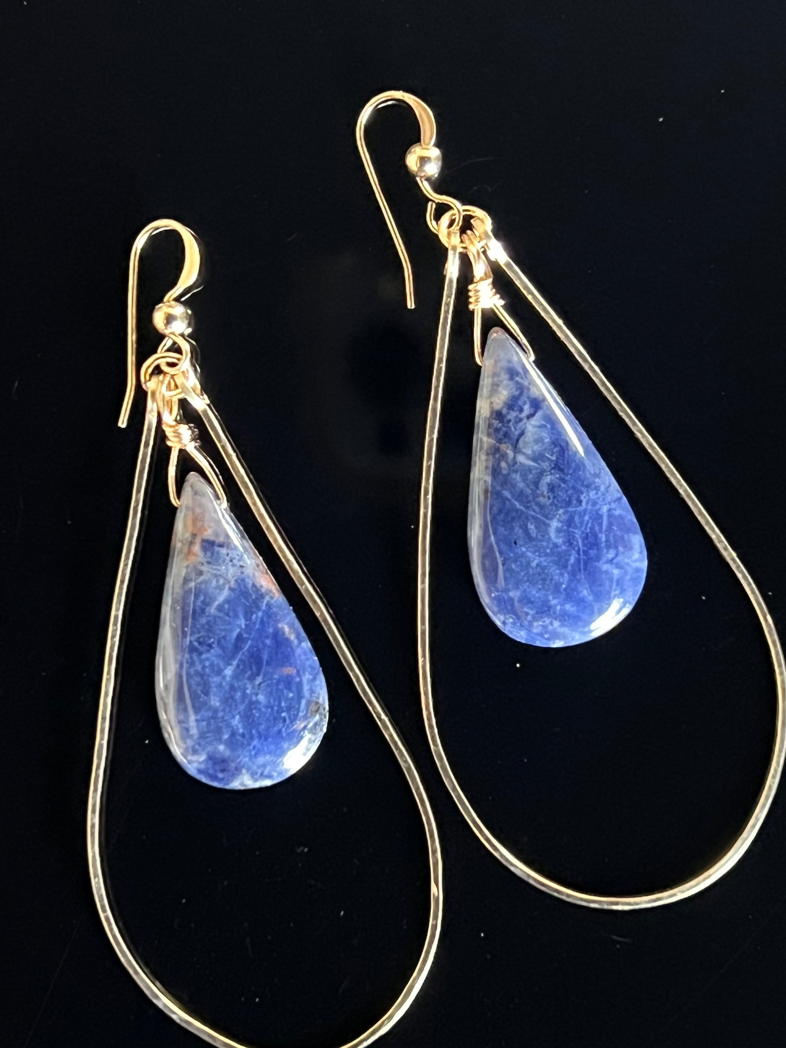 Gold teardrop earrings featuring blue sodalite gemstone pendants on a black background