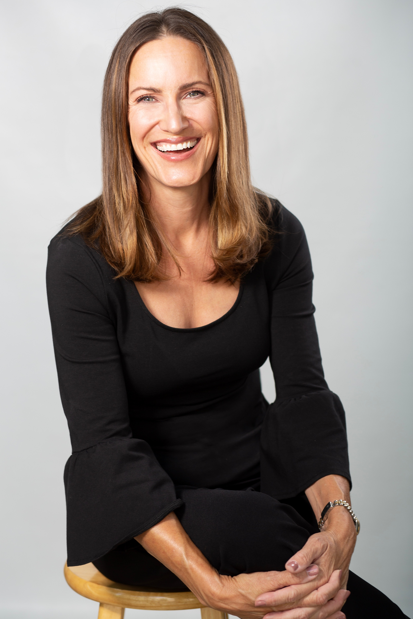 Smiling woman in black top seated on stool with relaxed posture against light background