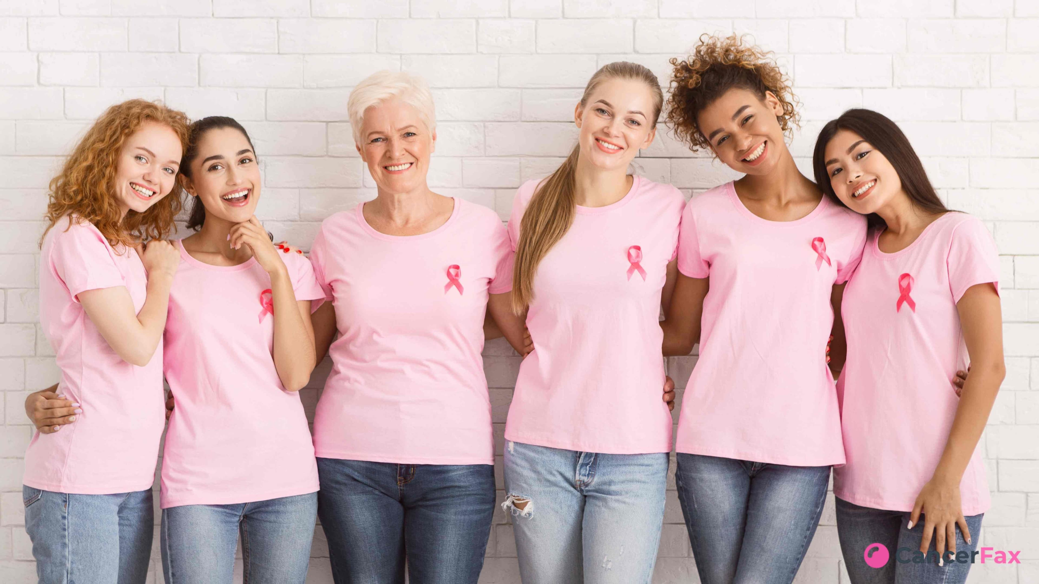 Six women in pink shirts stand together, smiling against a white brick wall