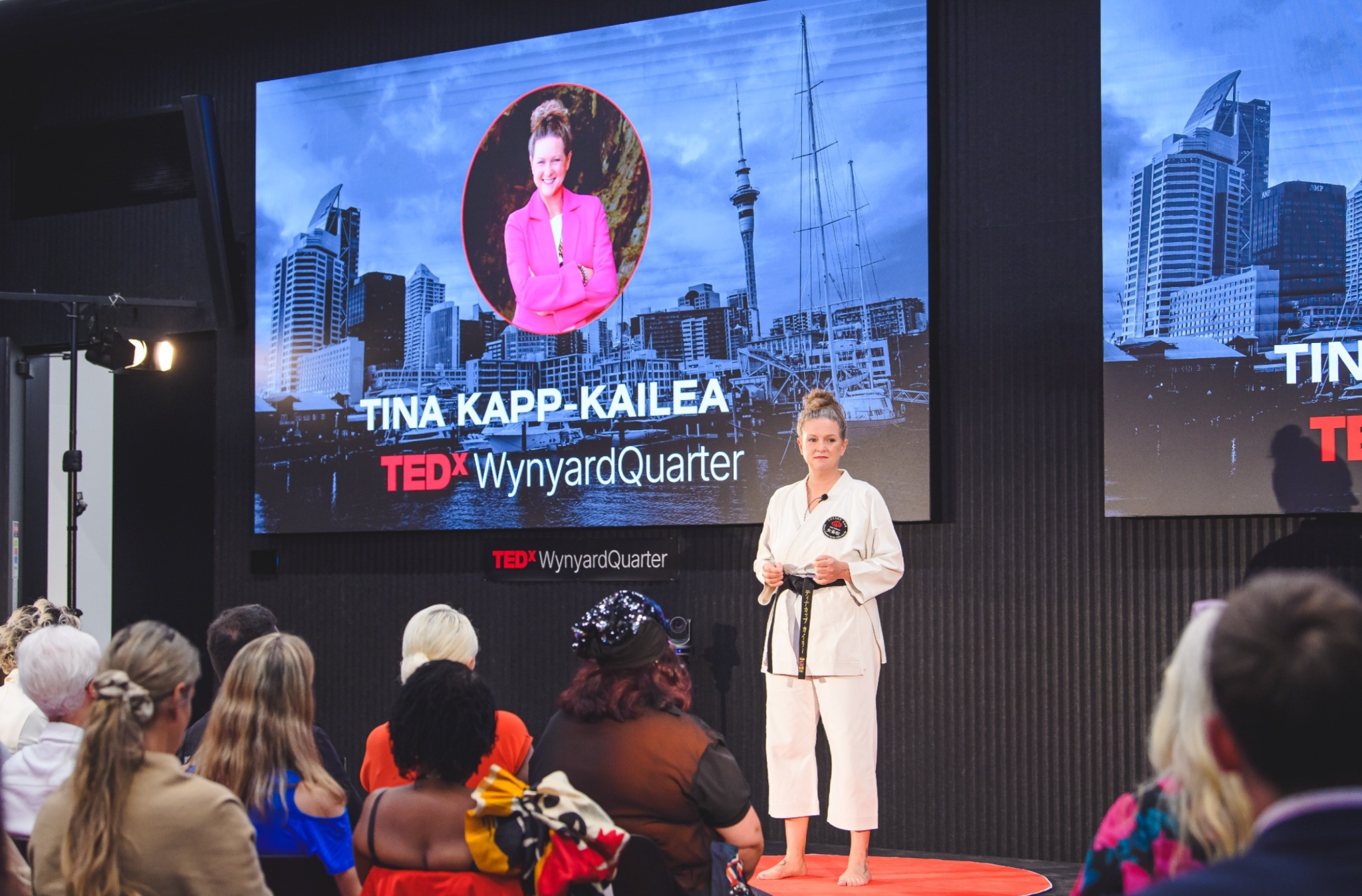 Speaker in martial arts attire presenting at a TEDx event with cityscape backdrop