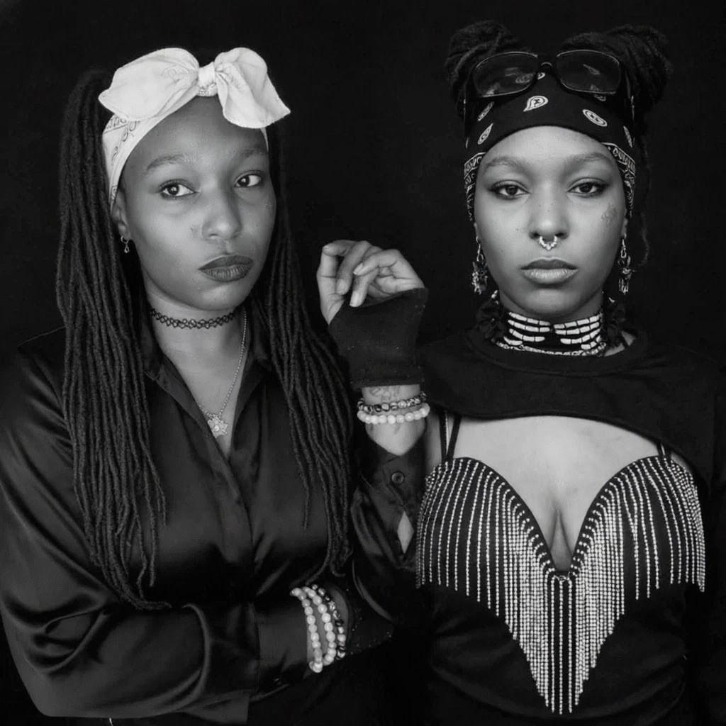 Two women in stylish black outfits pose fiercely against a dark background