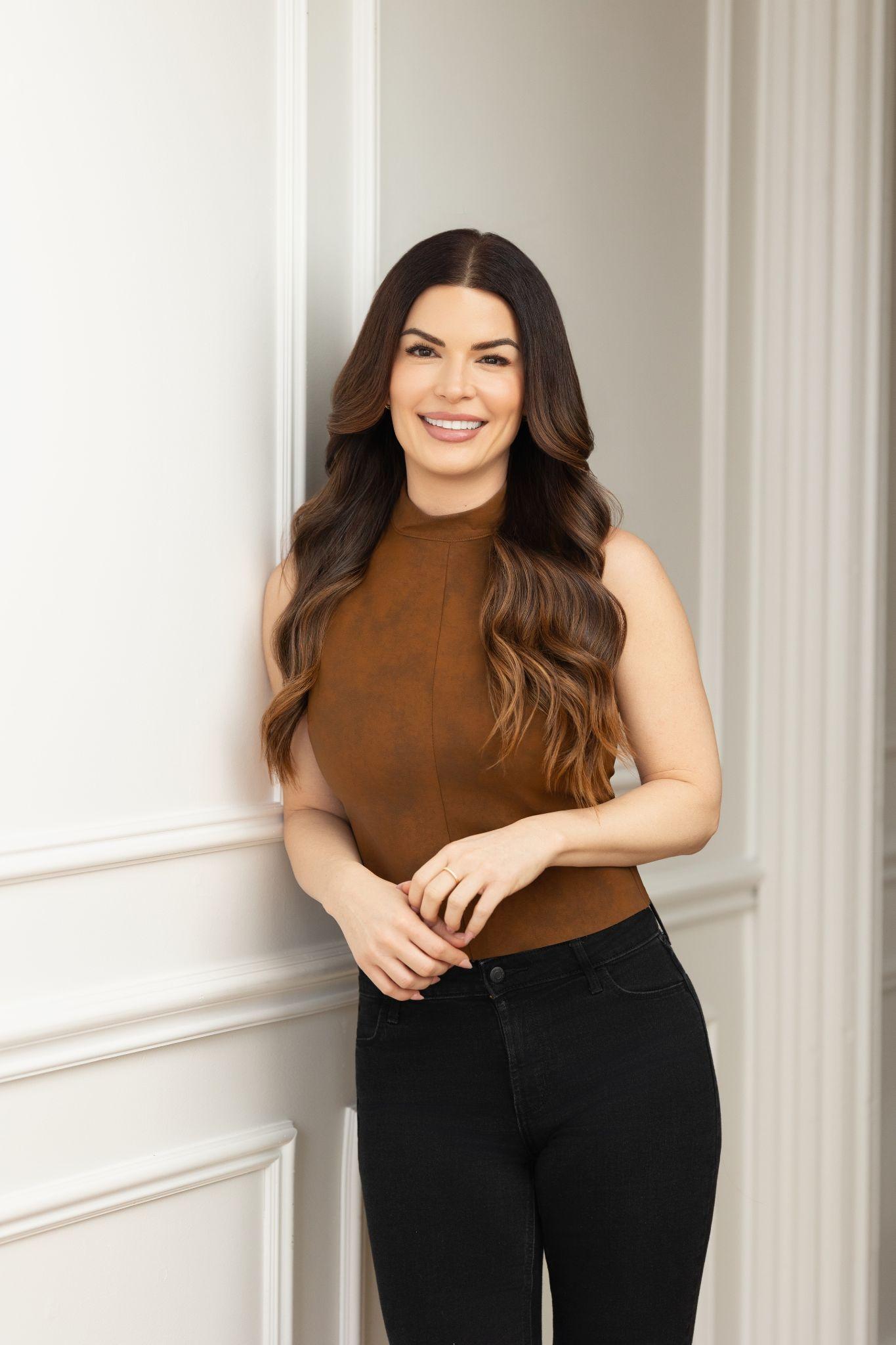 A woman with long brown hair poses smiling against a white wall.