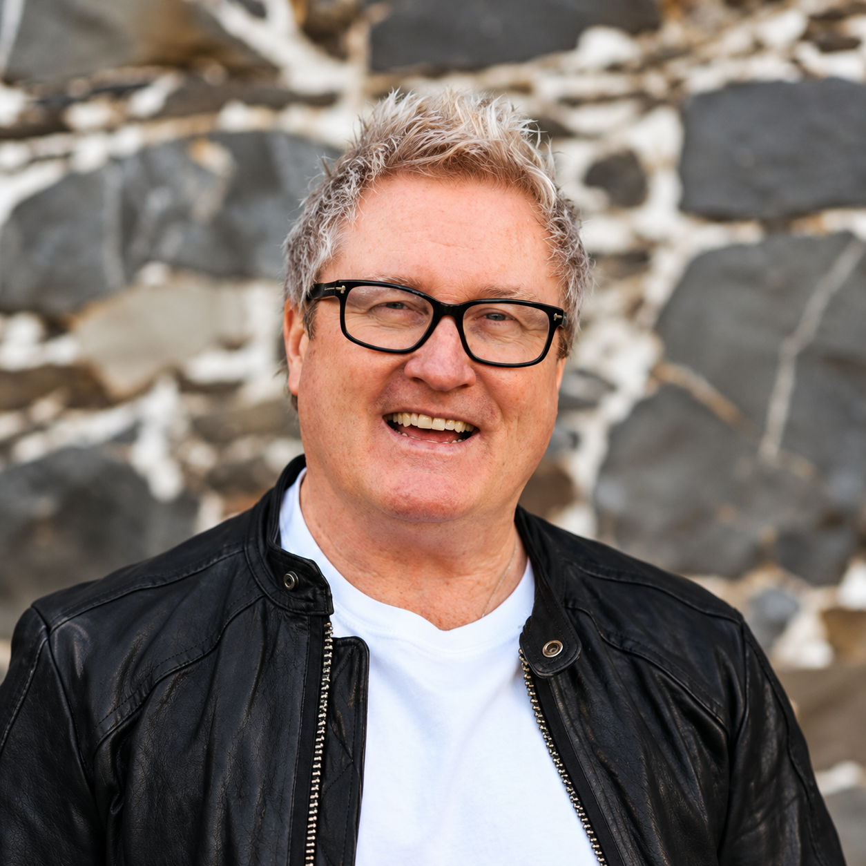 Smiling man with glasses wearing a black leather jacket against a stone wall background