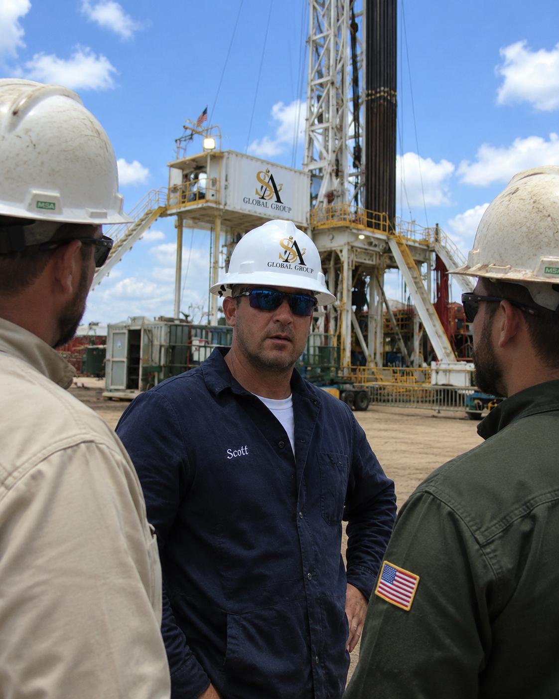 Three workers in hard hats discuss operations near a drilling rig in a field