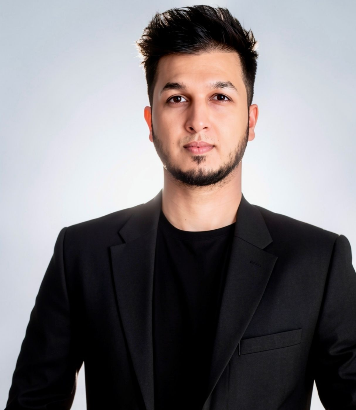 A young man in a black suit jacket poses confidently against a light background