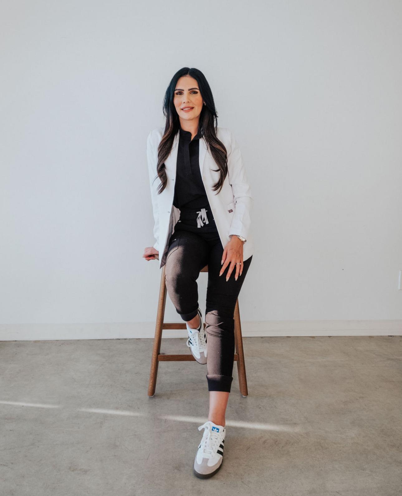 Woman in a white blazer and black outfit sitting on a wooden stool