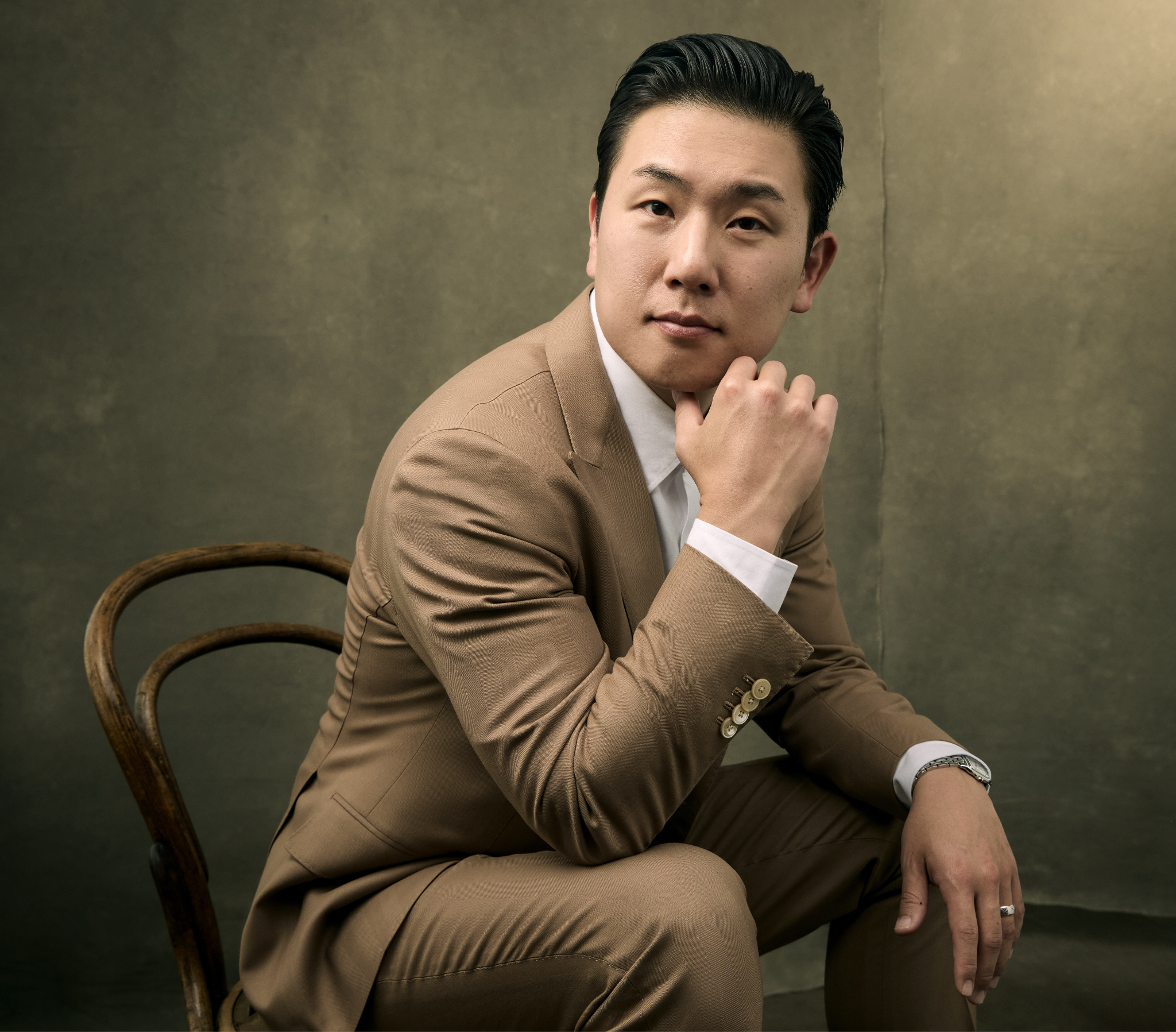 Man in a tan suit sits on a chair, thoughtfully posing with hand on chin