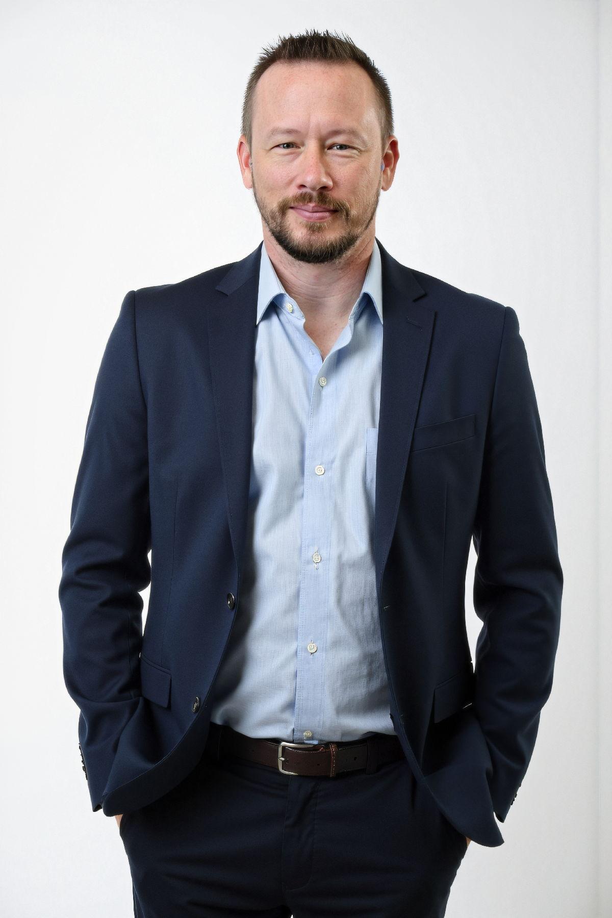 Man in a navy suit stands confidently with hands in pockets against a plain background