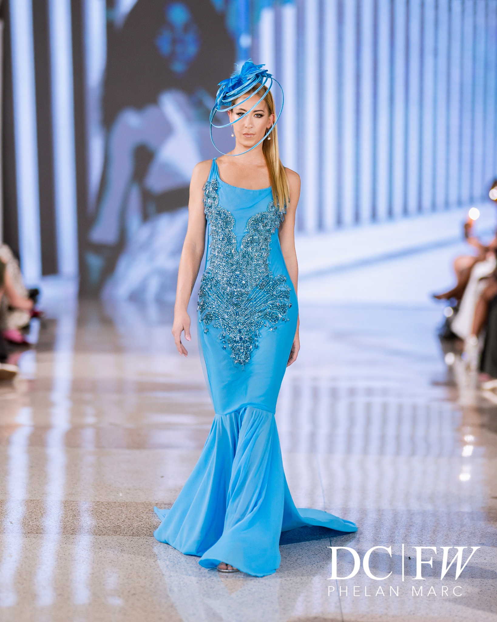 Model walks the runway in a blue gown with intricate embellishments and a unique hat