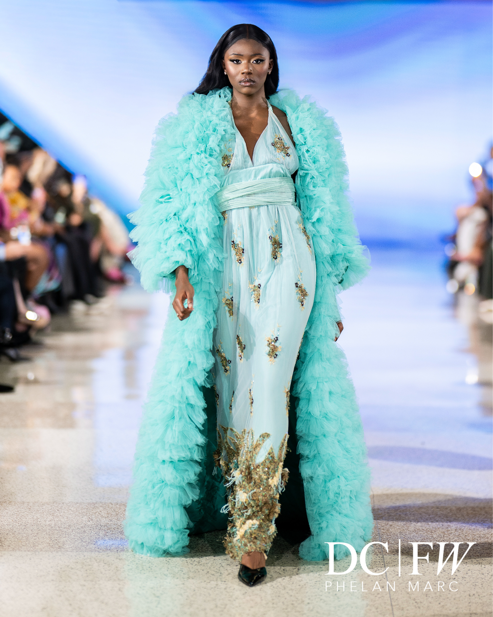 Model wearing a flowing teal gown with gold embellishments, styled with a matching fluffy coat.