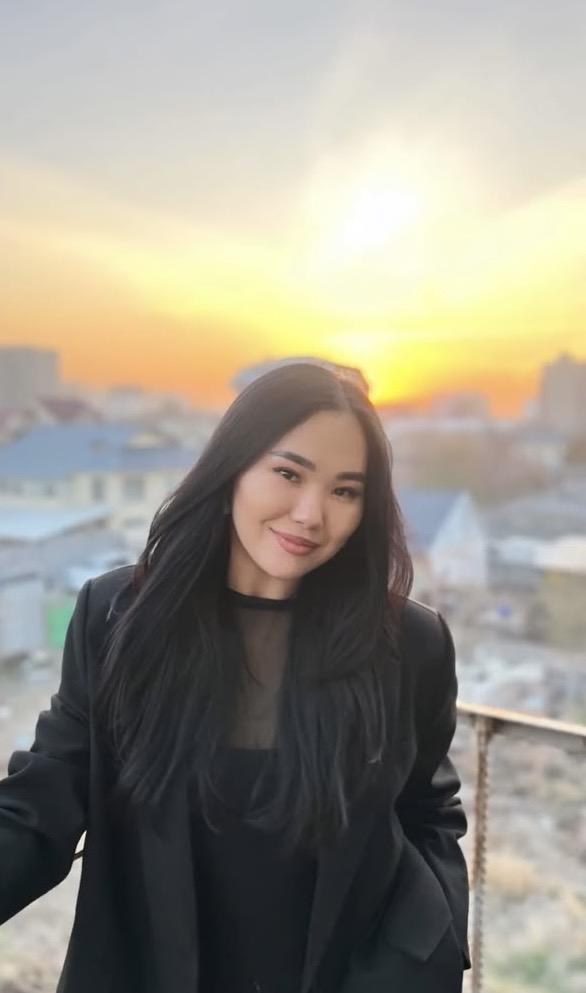 A woman with long black hair smiling against a sunset backdrop in an urban setting