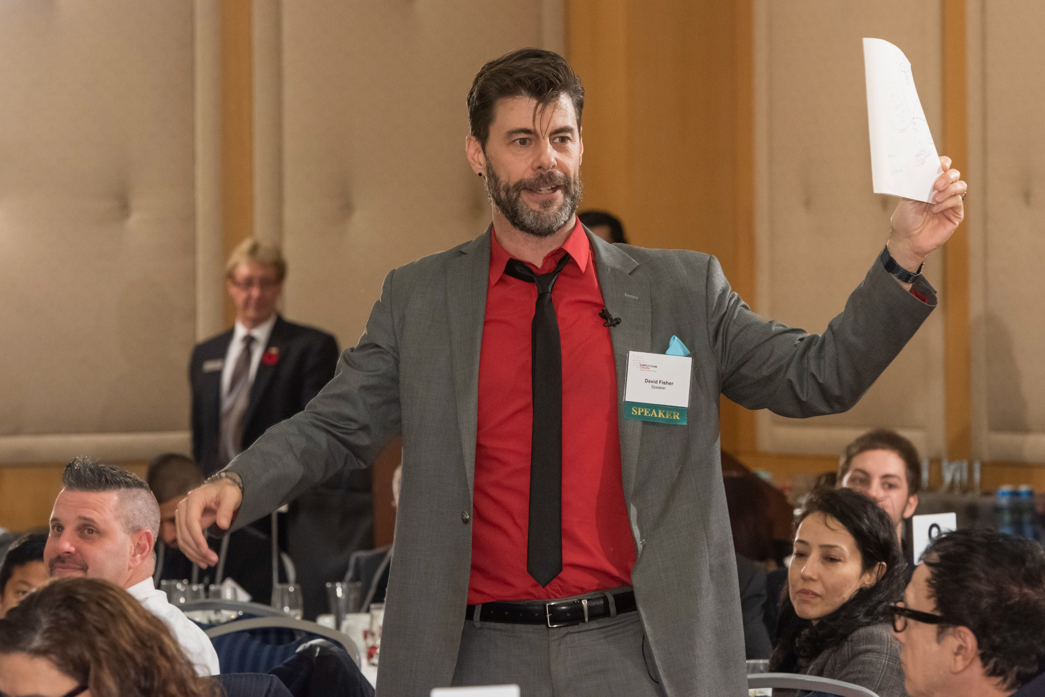 Man in gray suit holds paper while speaking at an event with attendees in view